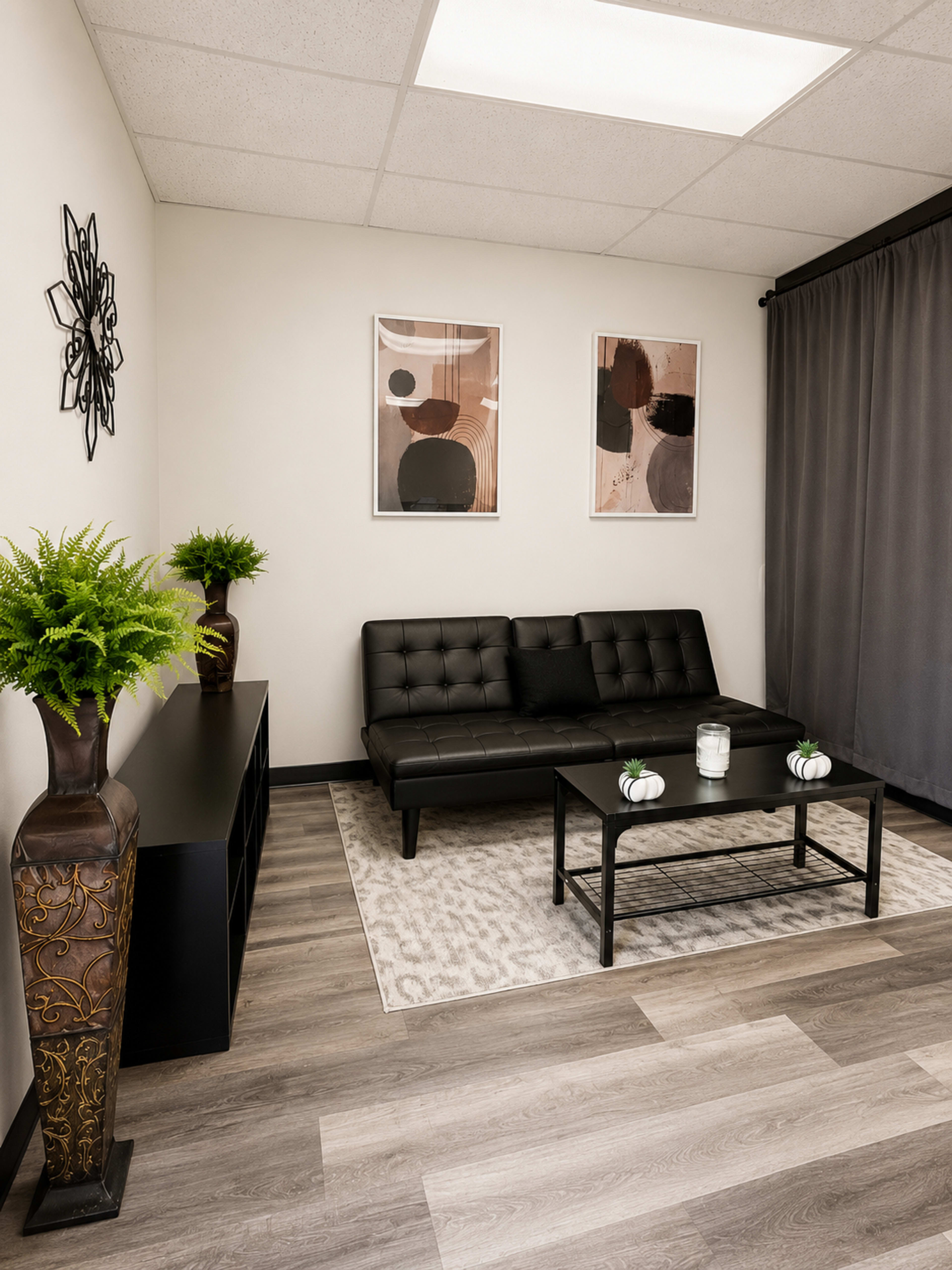 The image shows a modern lounge area featuring a black sofa, a low black coffee table, decorative plants, and framed abstract art on the walls.