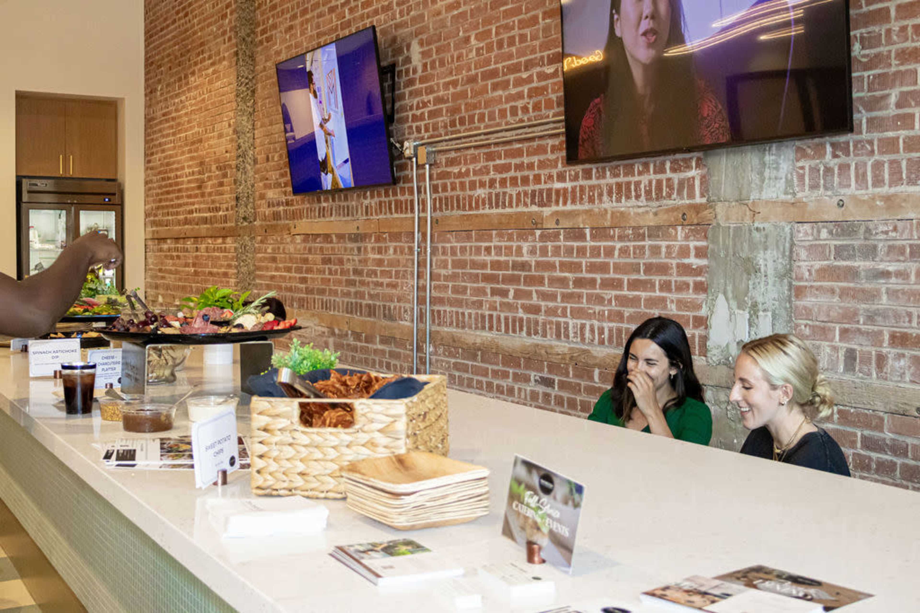 Two women are seated at a modern counter in a restaurant, with a display of food and drinks in front of them, while a staff member prepares food in the background.