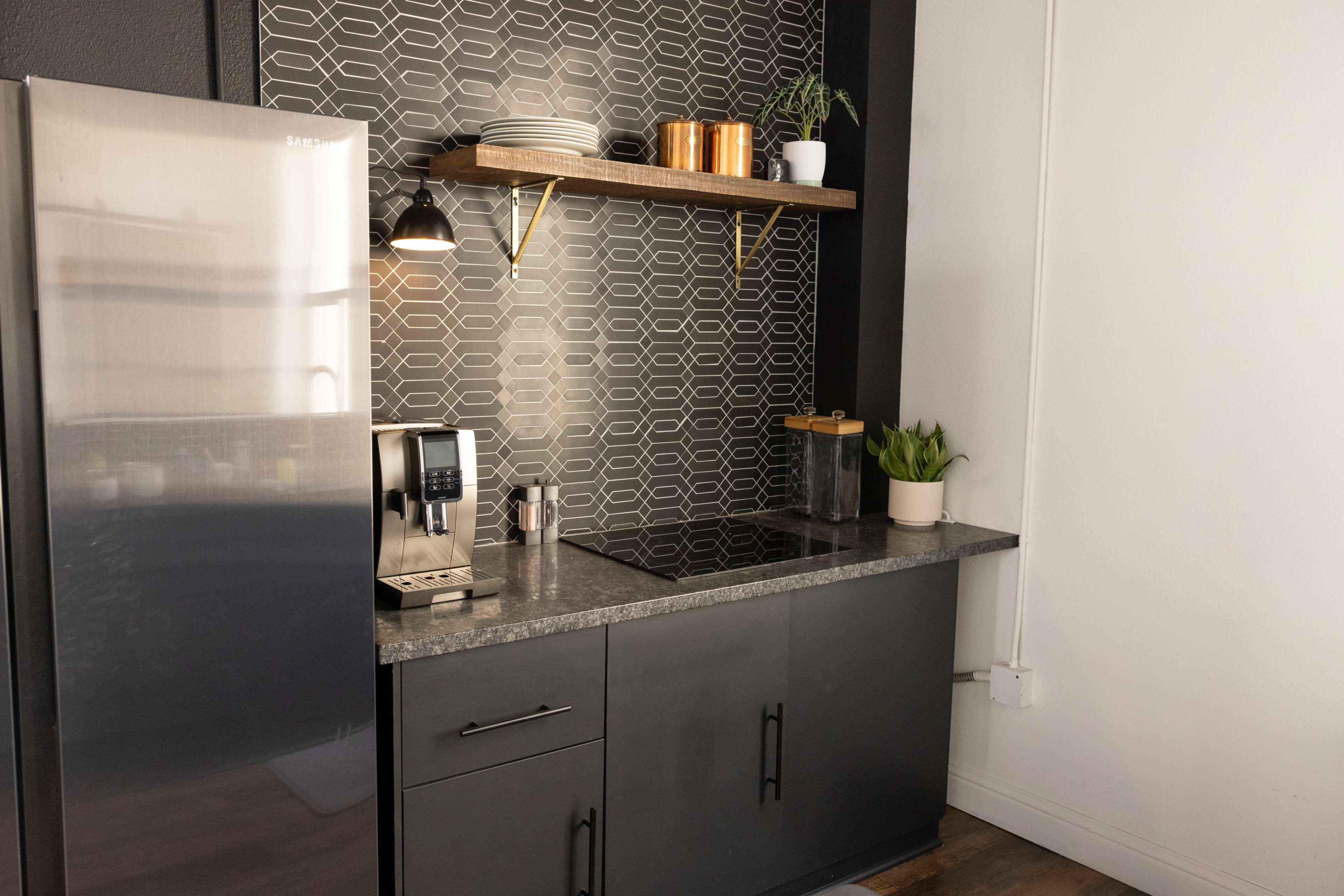 A modern kitchen corner features a stainless steel refrigerator, a sleek countertop with a coffee maker, and open shelving displaying plates and plant decor against a patterned backsplash.