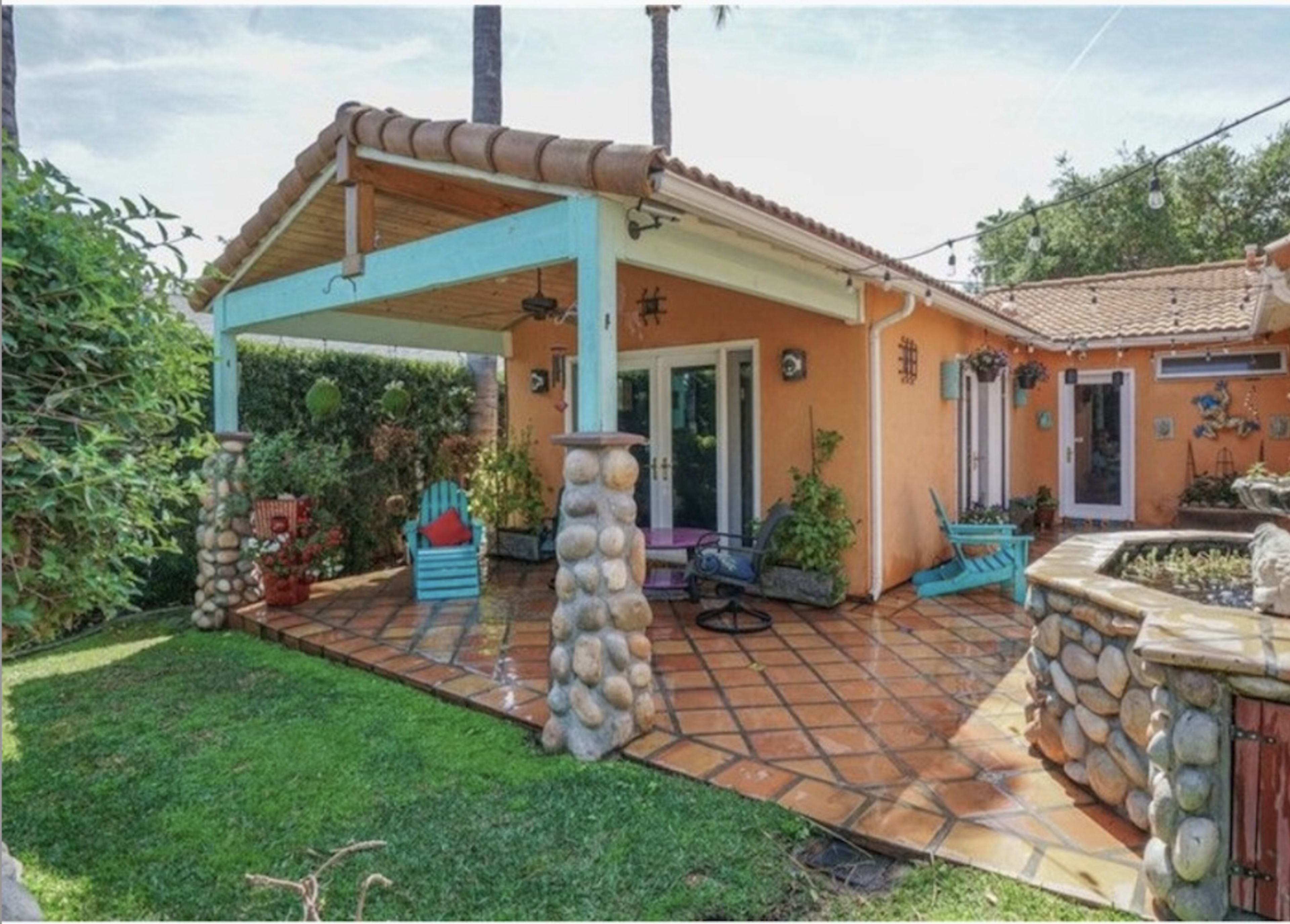 The image shows a colorful orange house with a stone patio, surrounded by greenery and featuring an outdoor seating area with chairs.