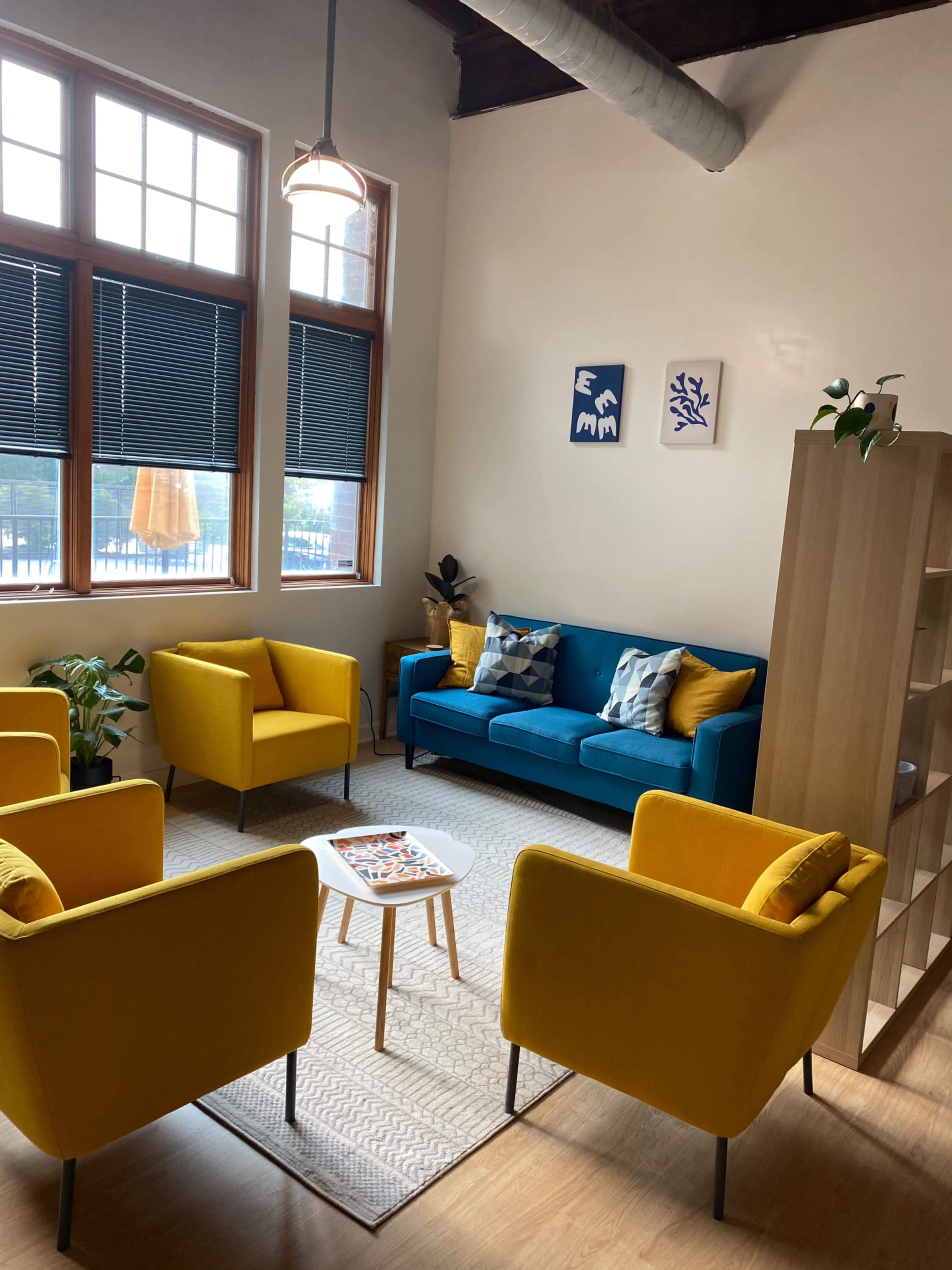 A cozy room with a blue sofa and four yellow chairs arranged around a small table, accented by decorative art on the walls and a plant in the corner.