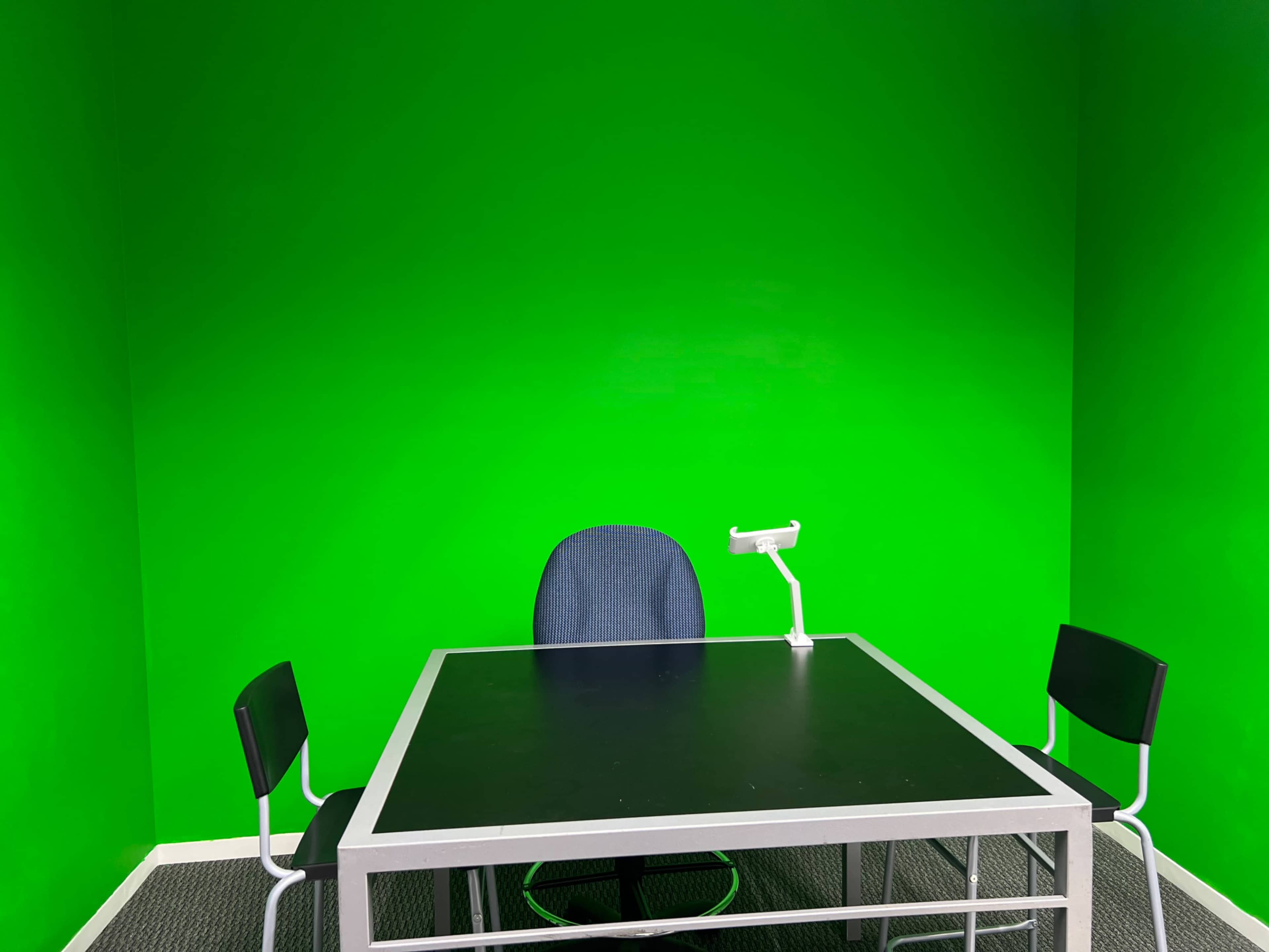 The image shows a room with a bright green wall, a black table, a blue office chair, and two black chairs.