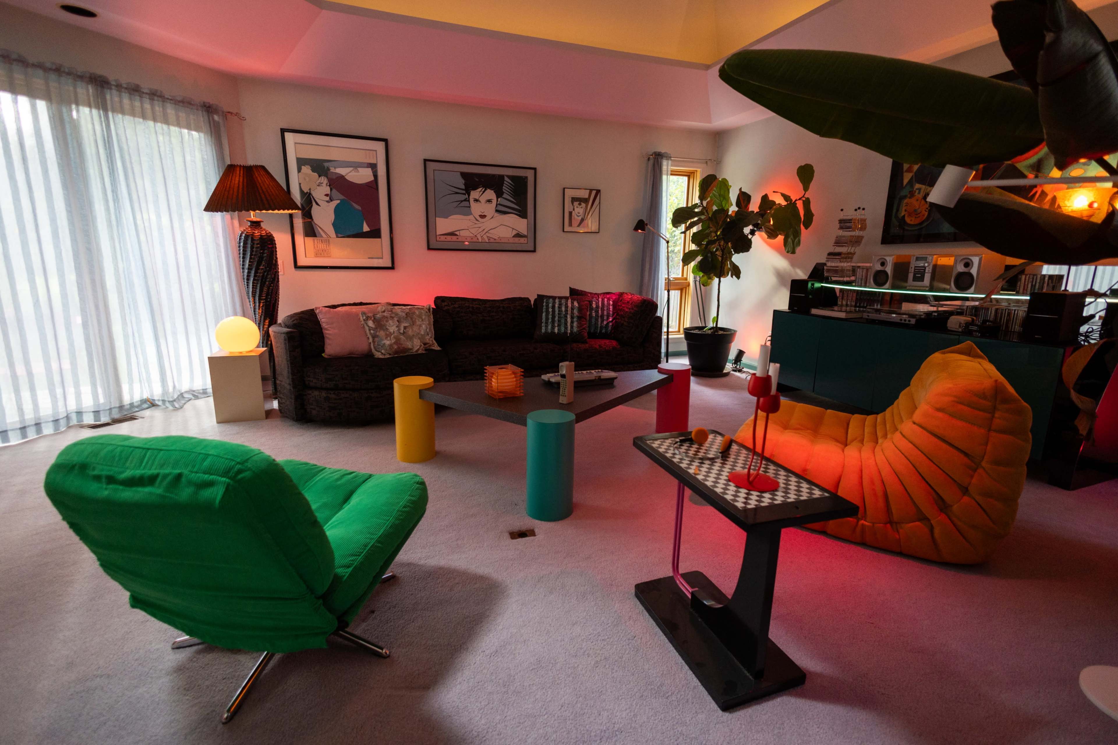 A modern living room features a dark sofa, colorful seating options, and a bright bar area, with large windows allowing natural light to filter in.