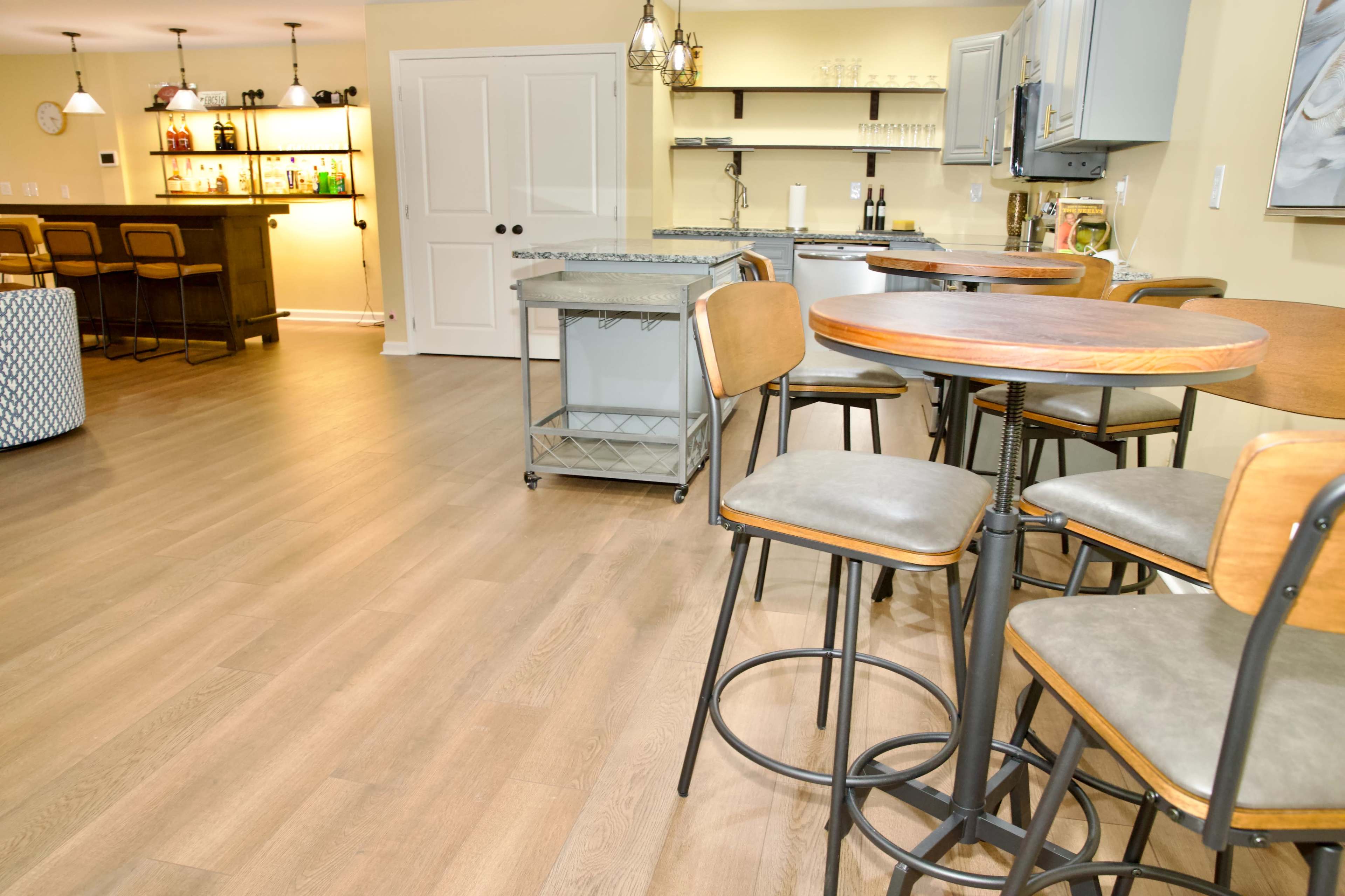 The image shows a spacious, well-lit room with modern bar tables and stools, a kitchen area in the background, and a bar counter with shelves.