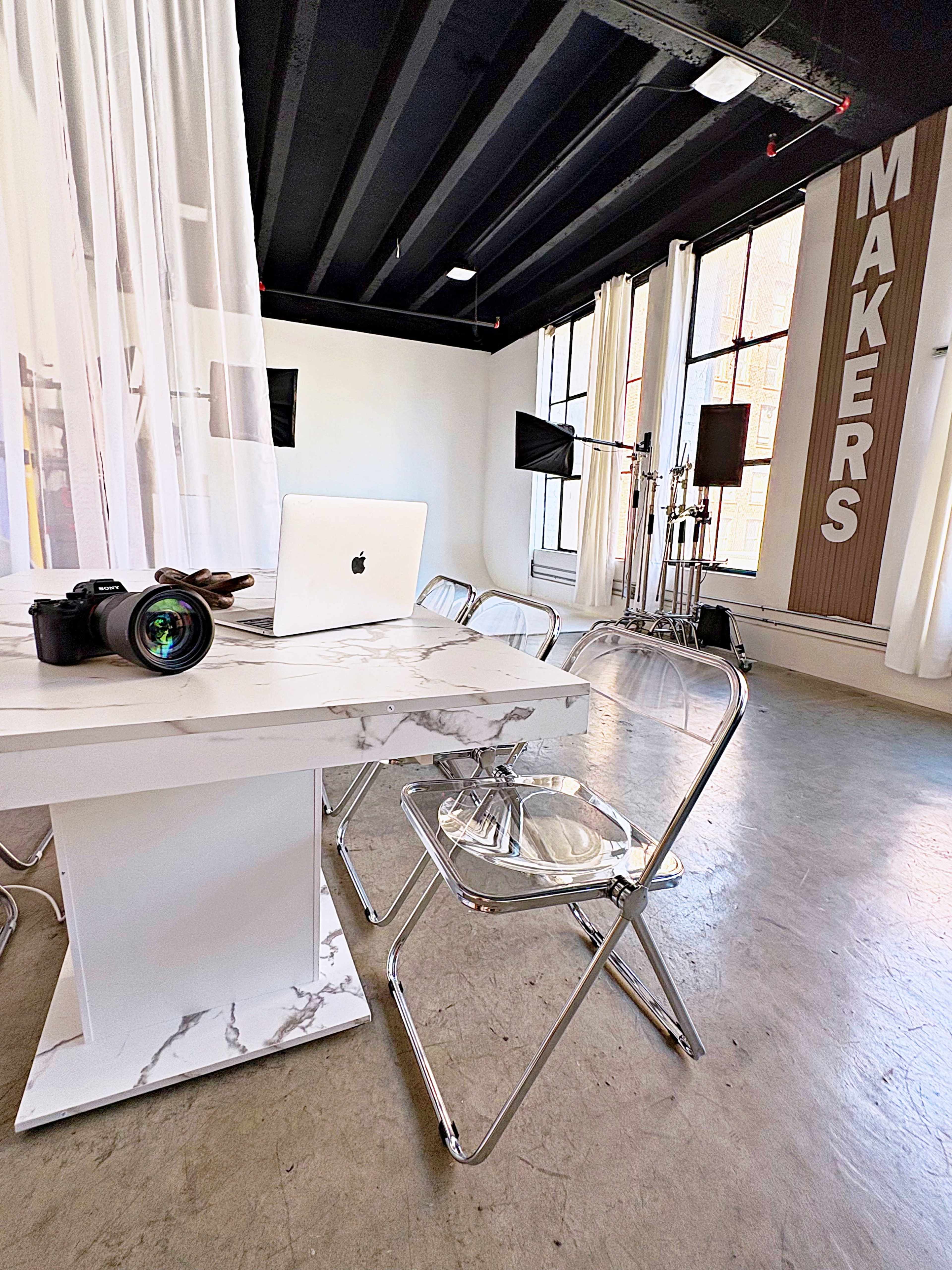 The image shows a modern workspace featuring a marble-top desk with a laptop and camera, surrounded by transparent chairs and large windows letting in natural light.