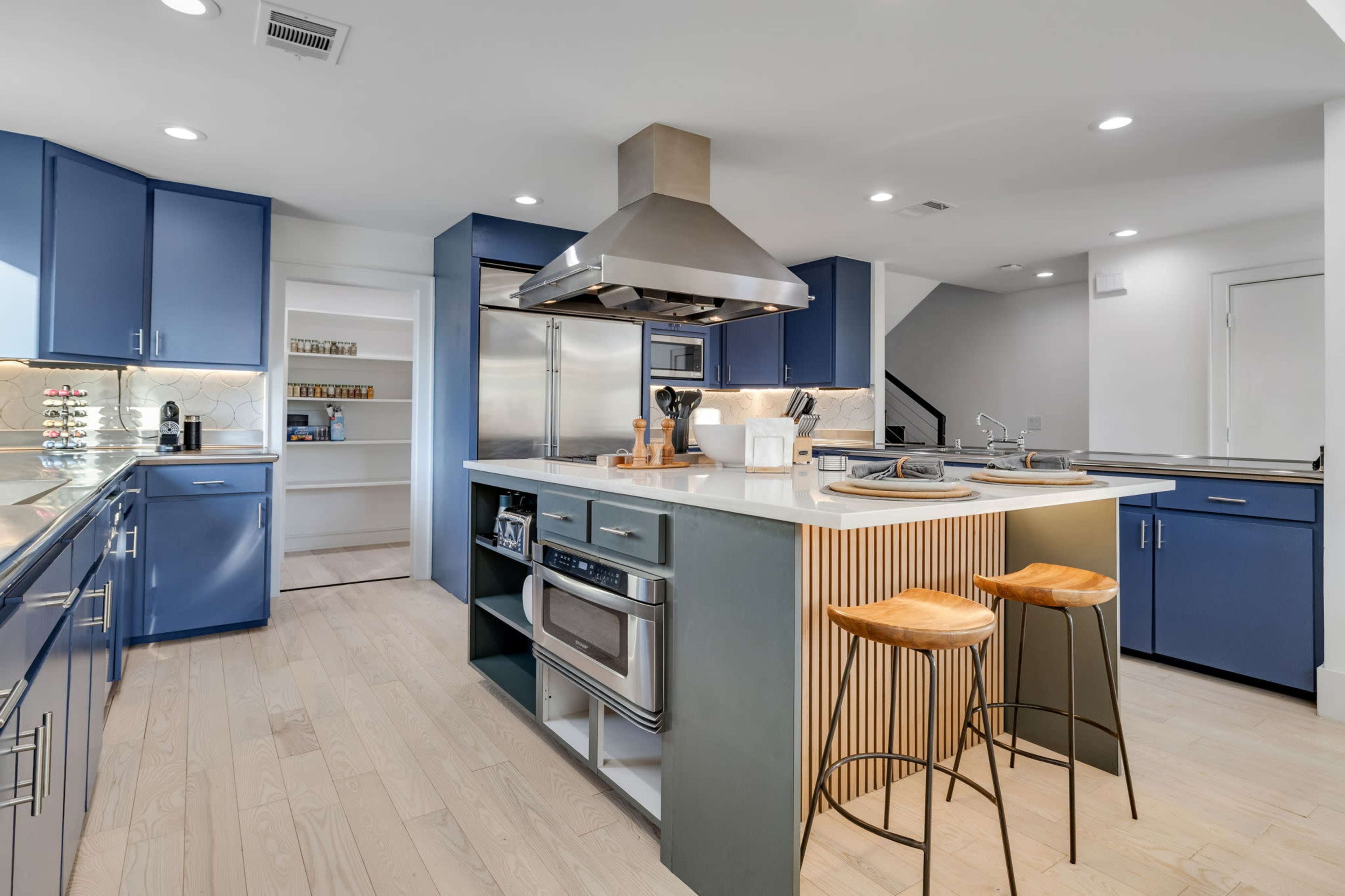 A modern kitchen features blue cabinetry, a central island with bar stools, stainless steel appliances, and ample natural light.