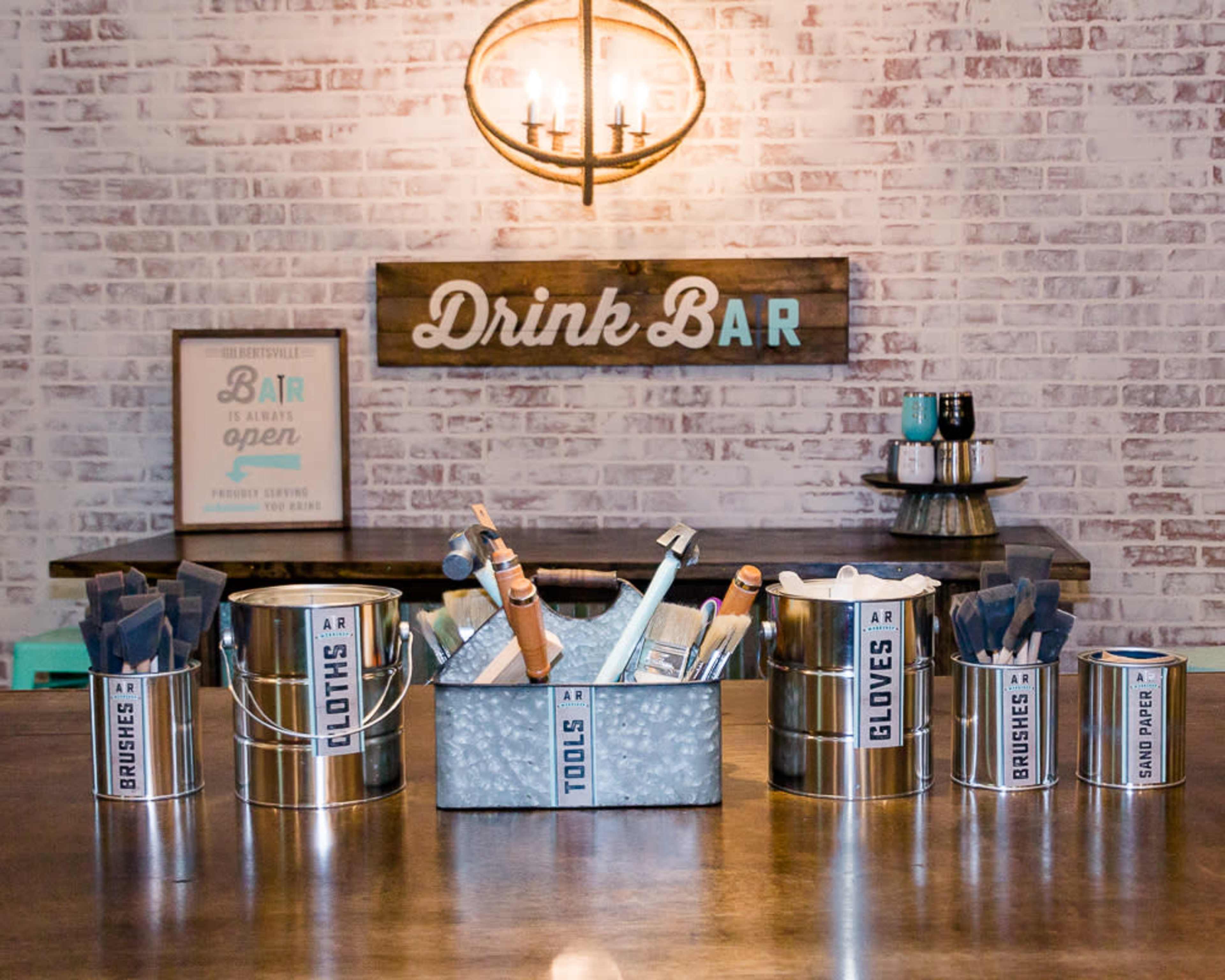 A bar setup with containers labeled "SHOES," "GLASSES," "BRUSHES," "TOOLS," and "SAND PAPER," arranged on a countertop under a rustic chandelier.