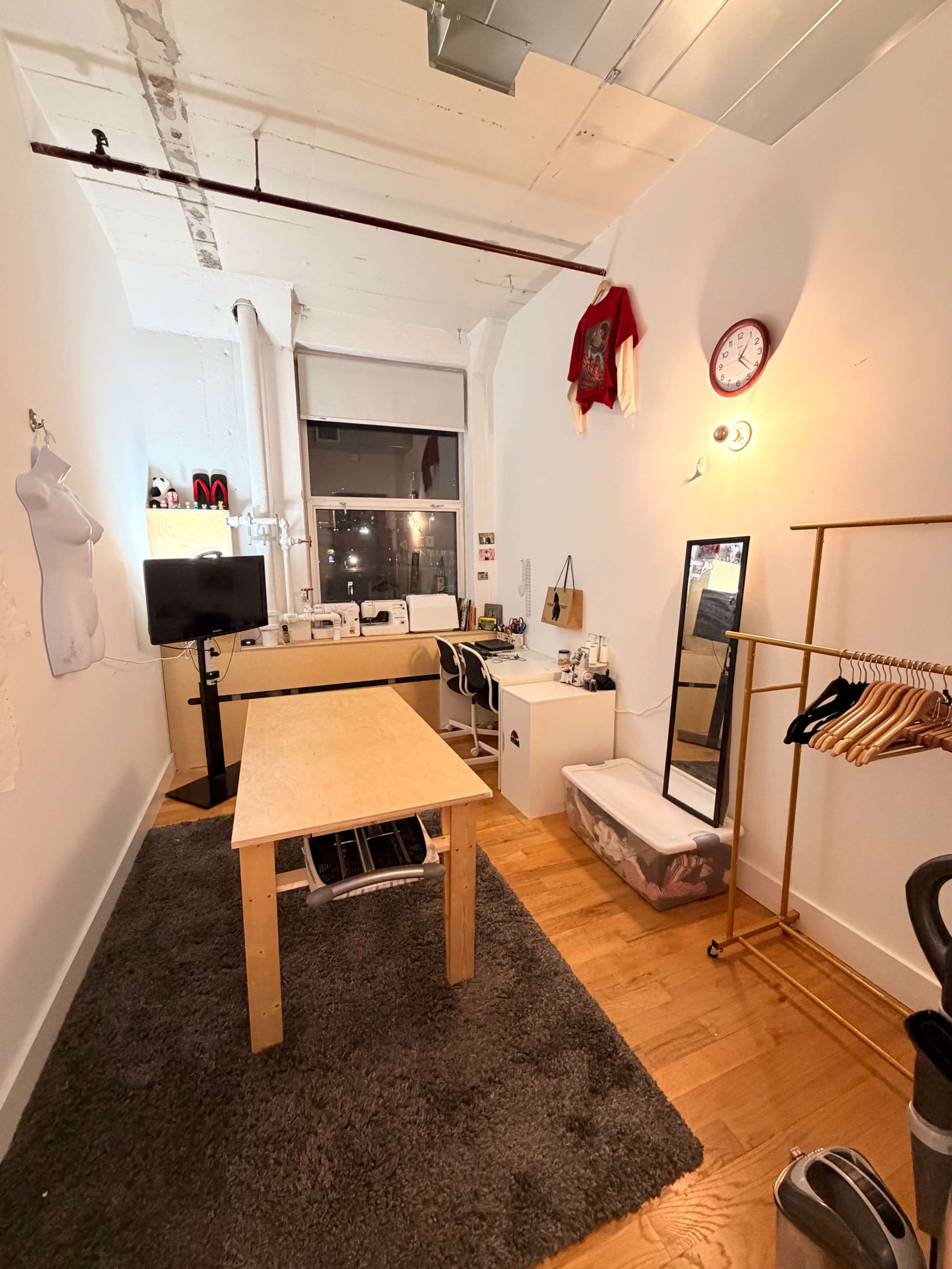 The image shows a small studio room with a wooden table at the center, a clothing rack on the right, and a workspace featuring a mirror and a television on the left.