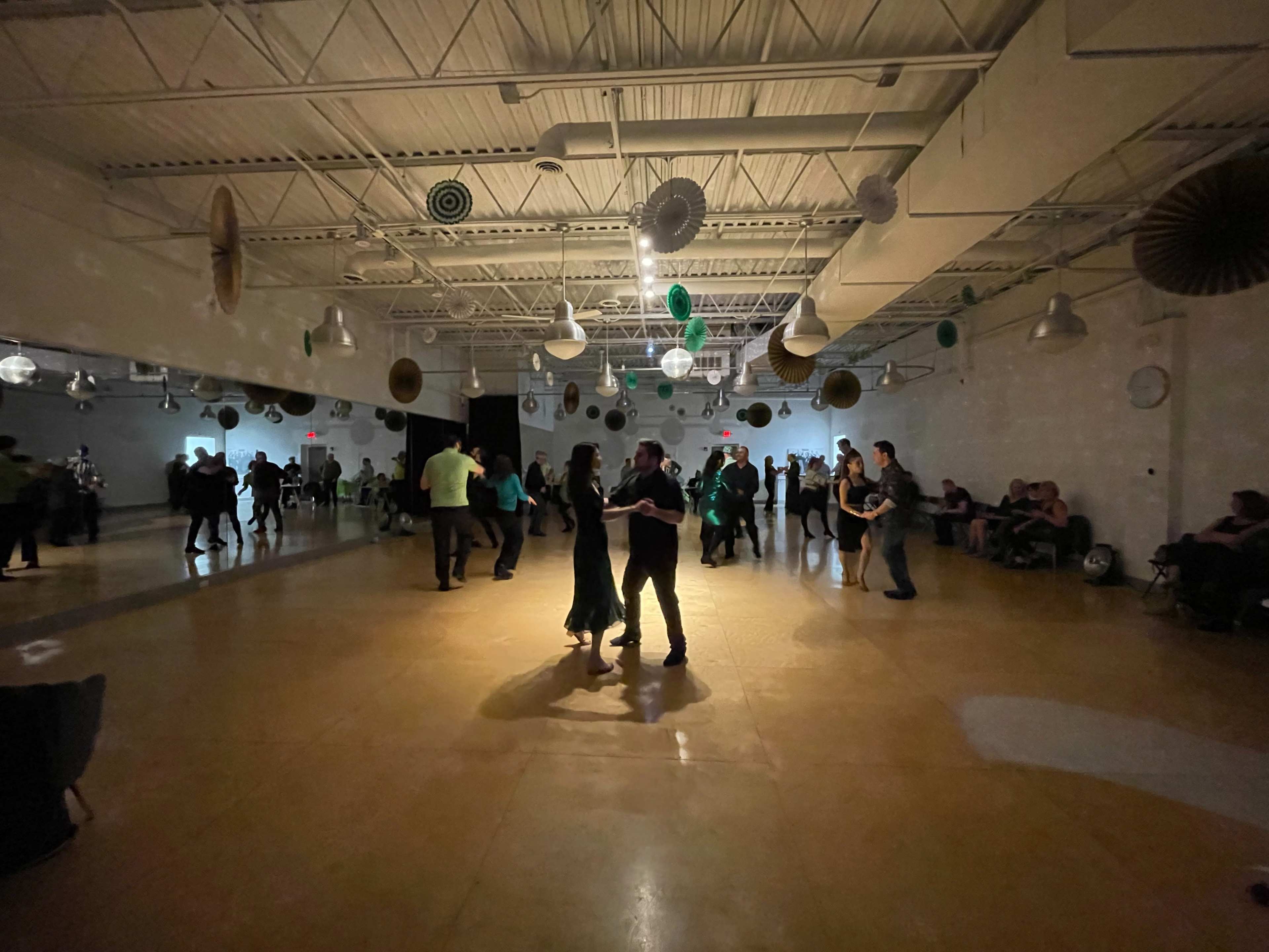 A spacious dance hall is filled with people dancing in pairs, while others sit along the walls under dim lighting.