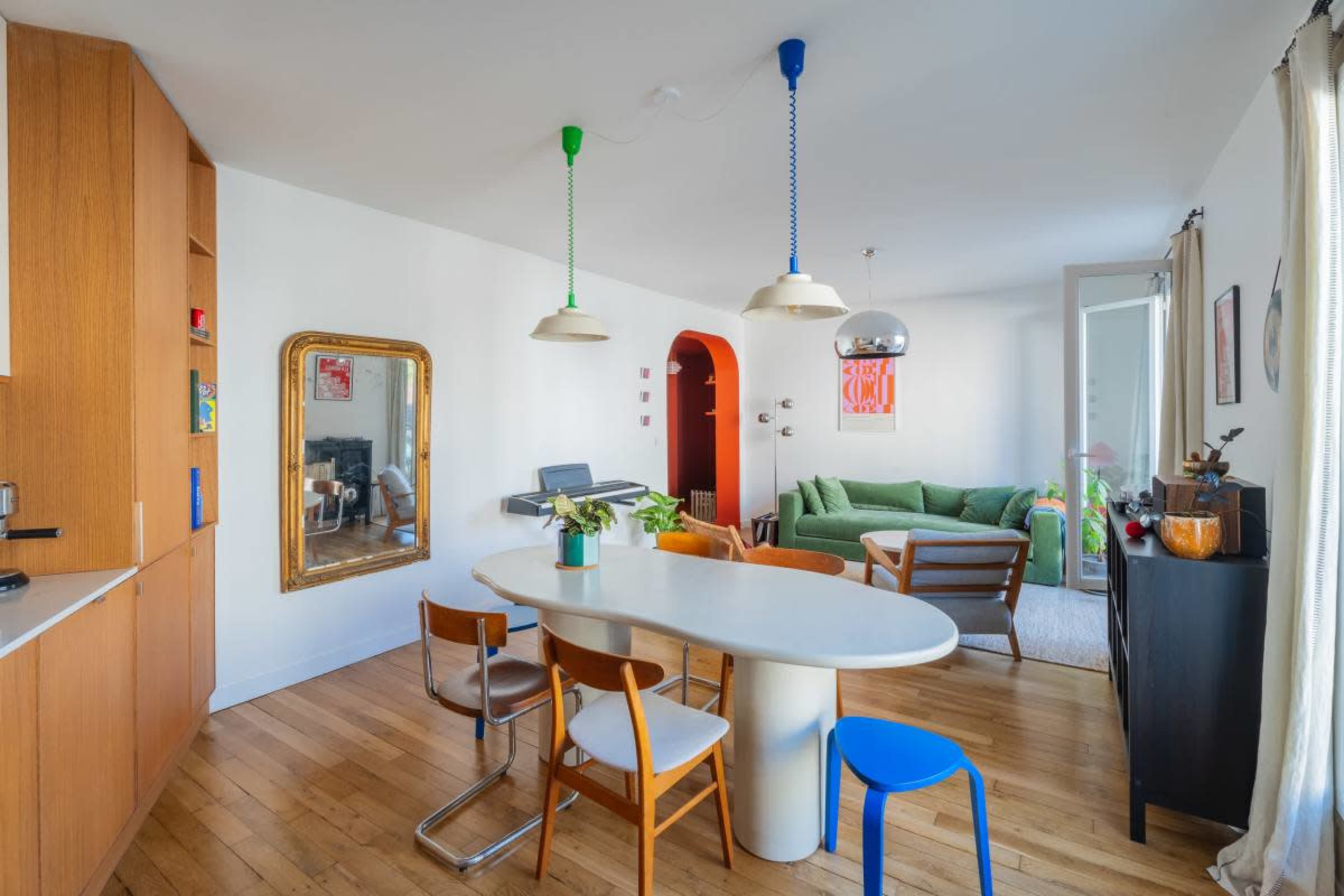 A bright and modern living space features a white dining table surrounded by wooden chairs, with an adjacent green sofa and a piano.