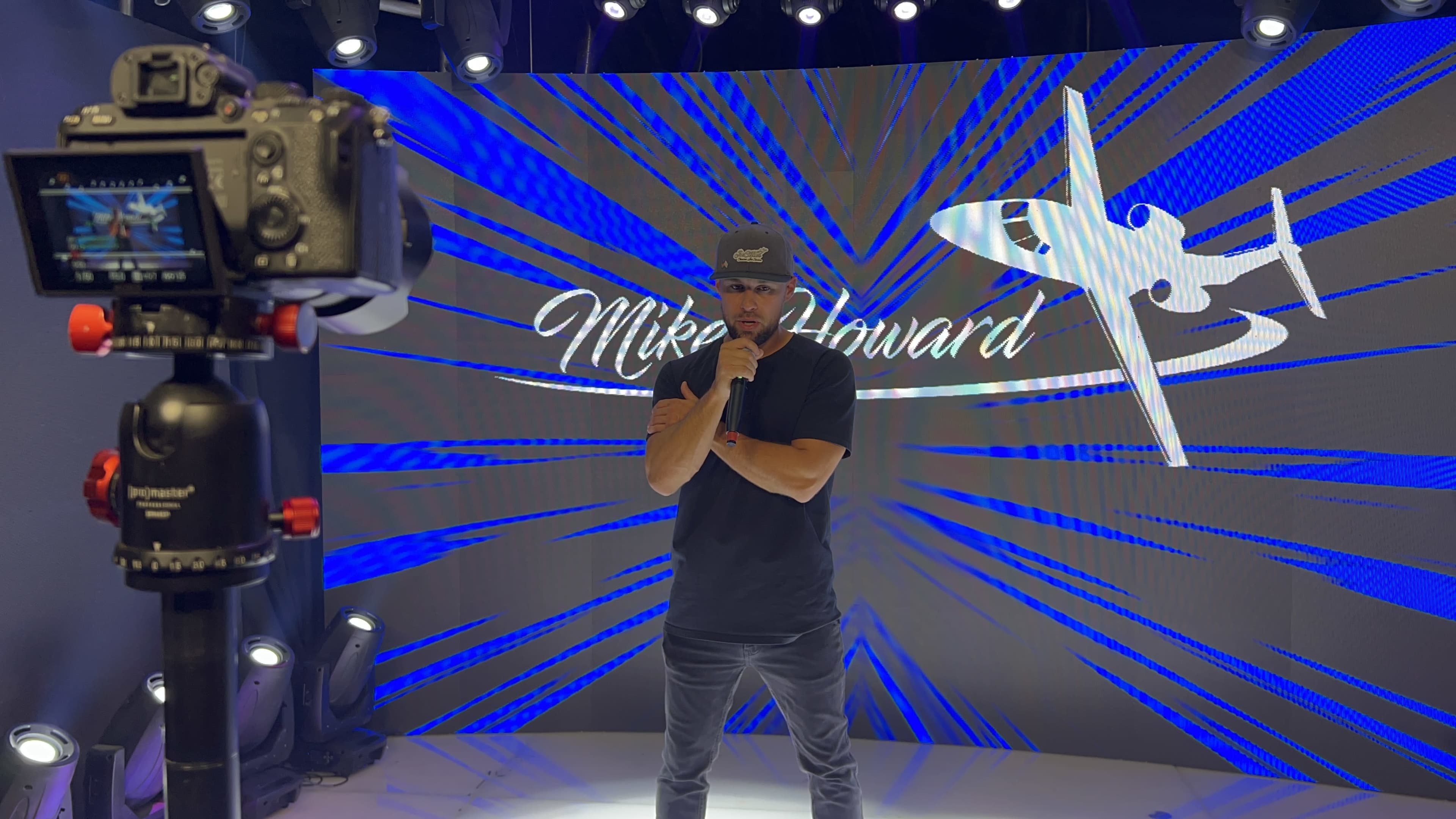 A man stands with his arms crossed in front of a large projection screen displaying the name "Mike Howard" and a graphic of an airplane.