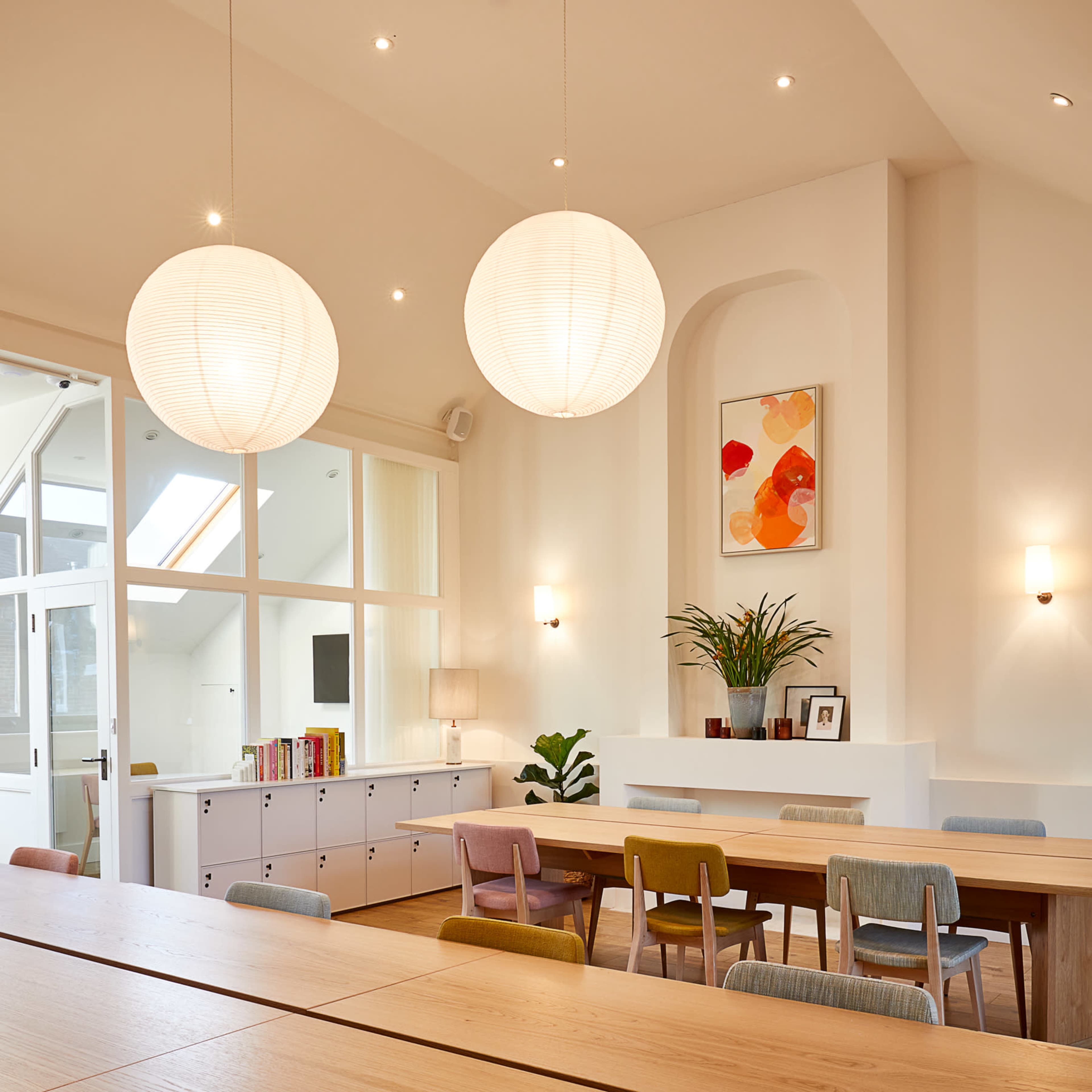 A well-lit, modern meeting room features two large paper lanterns, a long wooden table, and colorful chairs, with a white cabinet and artwork on the walls.