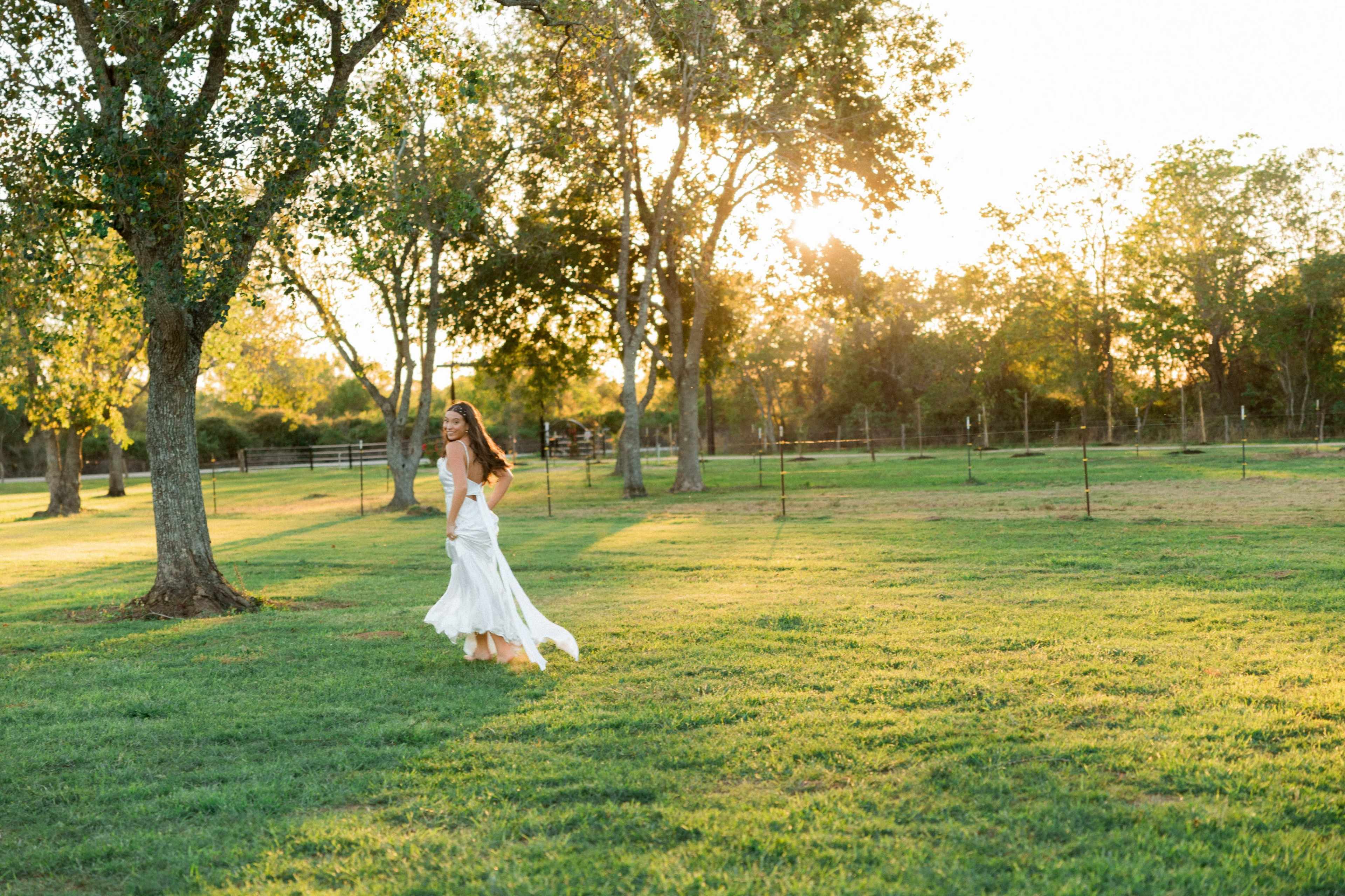 A woman in a white dress runs across a grassy field at sunset, surrounded by trees and a fenced area.