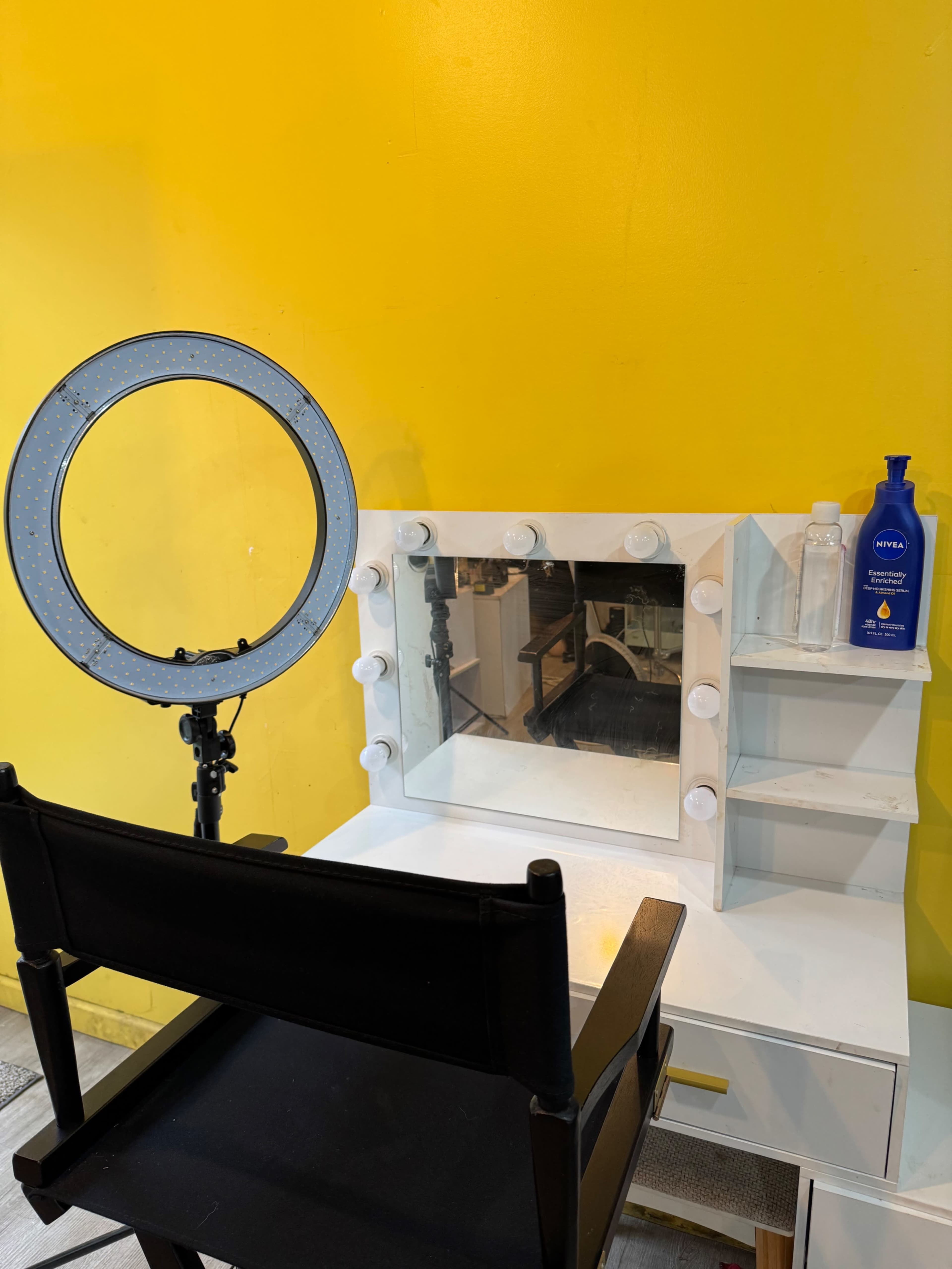 The image shows a makeup station with a mirror framed by light bulbs, a ring light, a black chair, and several cosmetic products on a yellow wall backdrop.