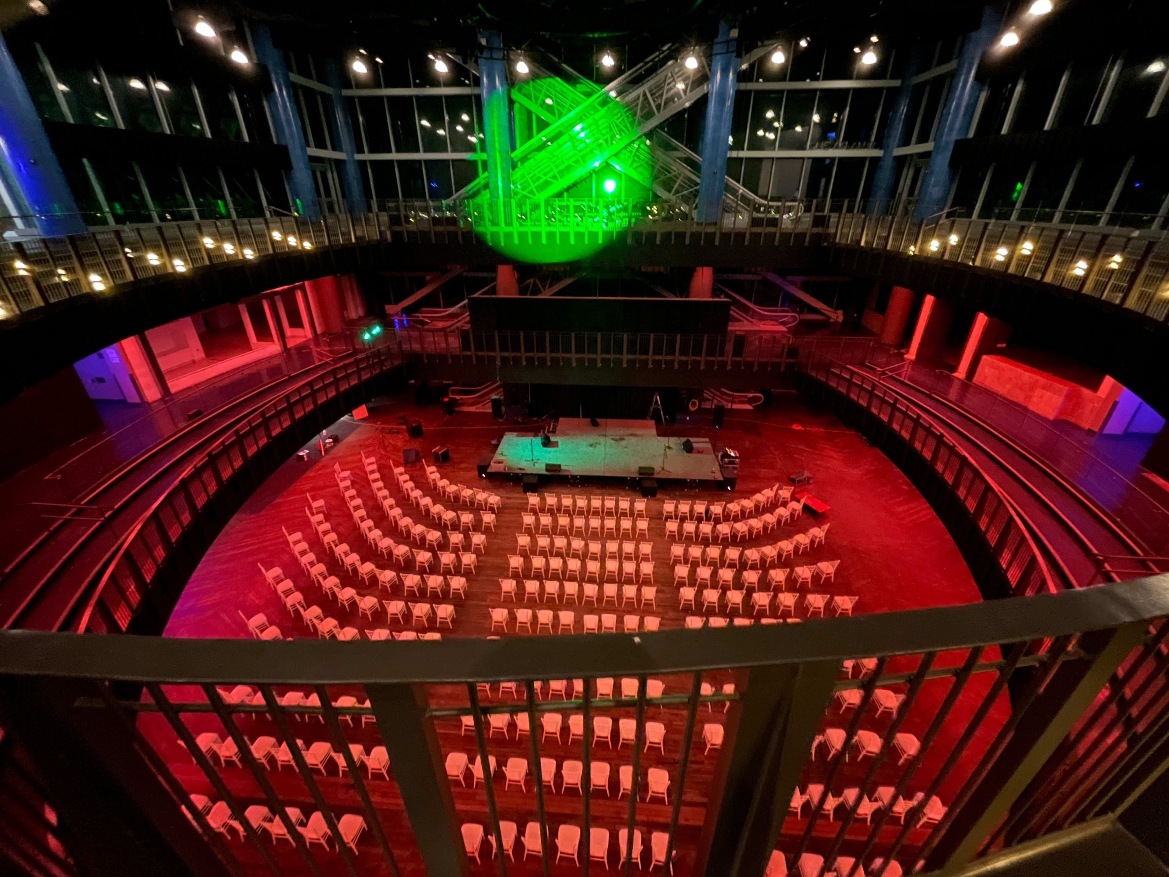 An empty theater-style venue with arranged white chairs surrounding a central stage, illuminated by colorful lighting effects.
