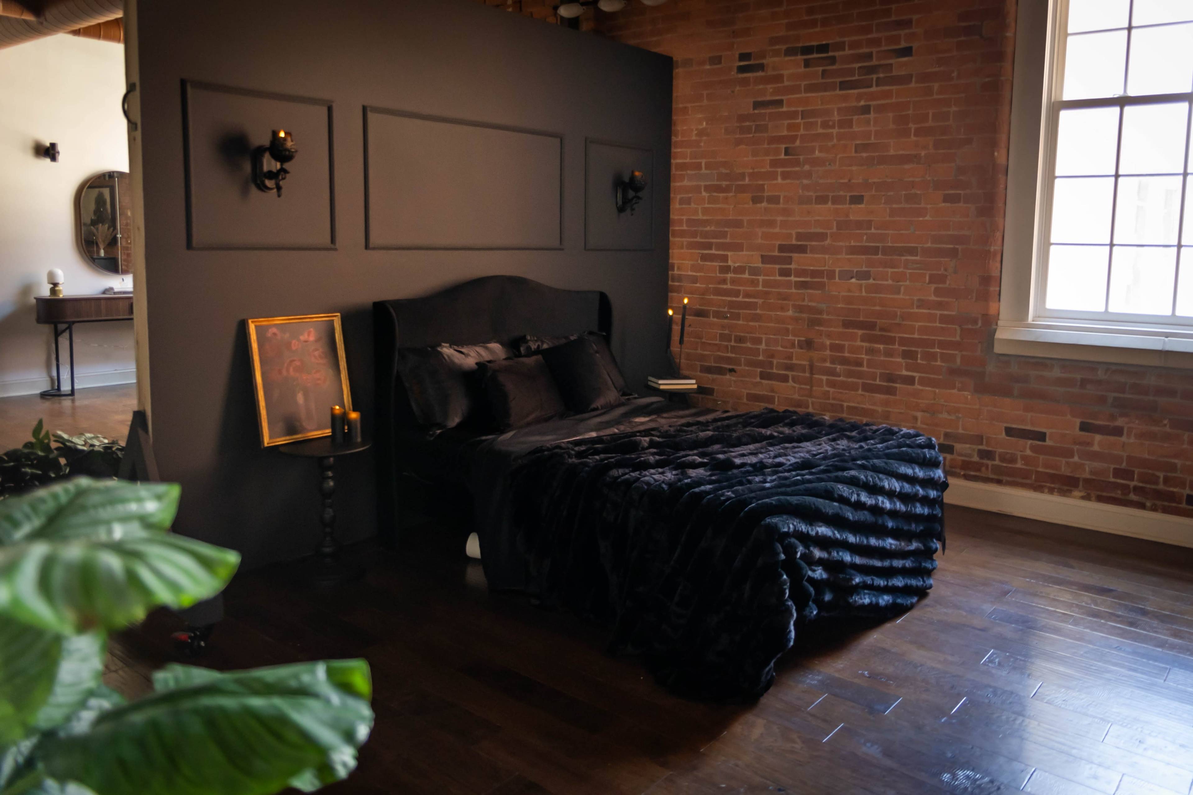 The image shows a stylish bedroom featuring a dark wall, a bed with a black fur blanket, and exposed brick accents.
