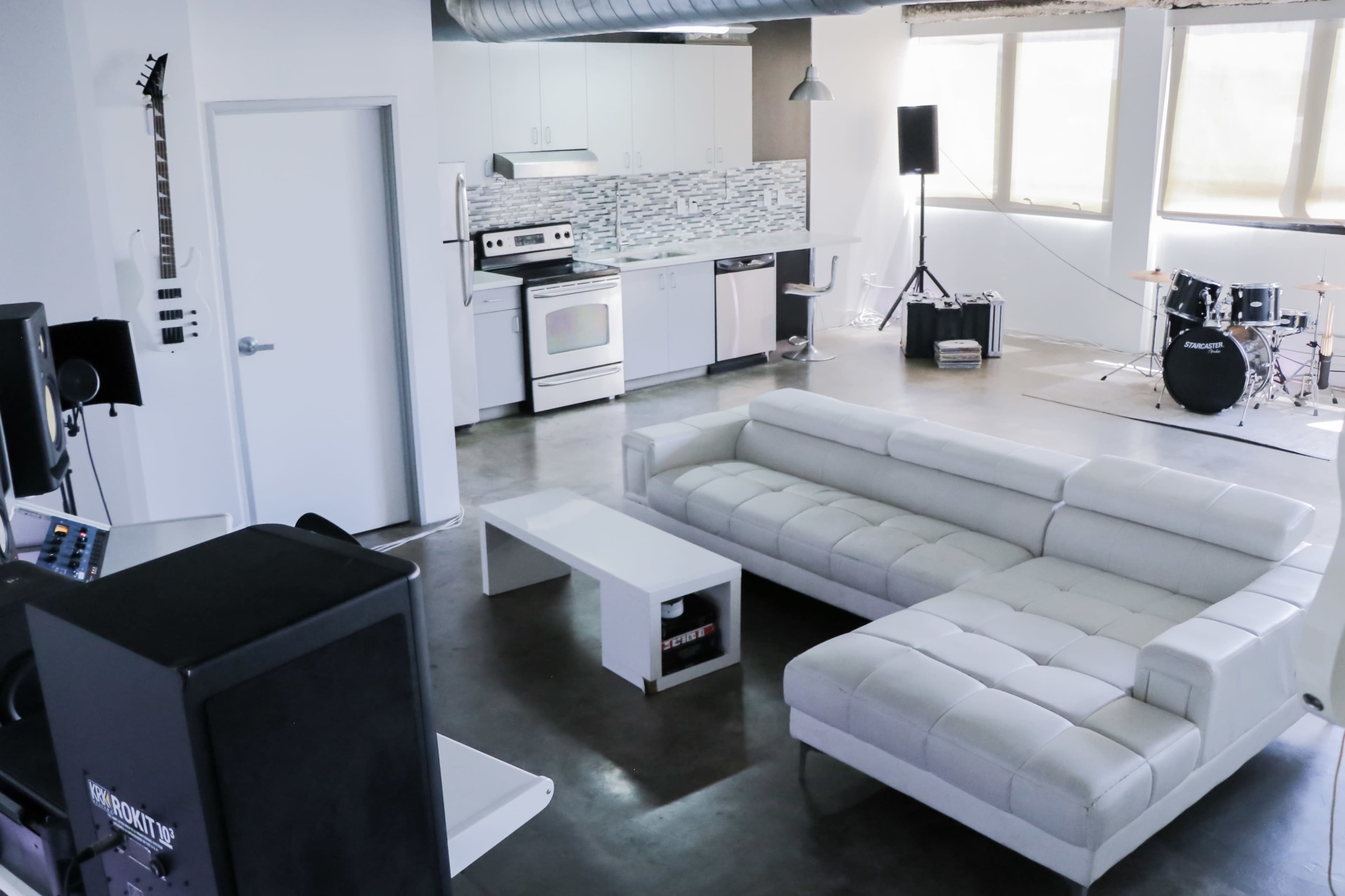 A modern living space featuring a white sectional sofa, a kitchen area with stainless steel appliances, and a small music setup with a drum kit and speakers.