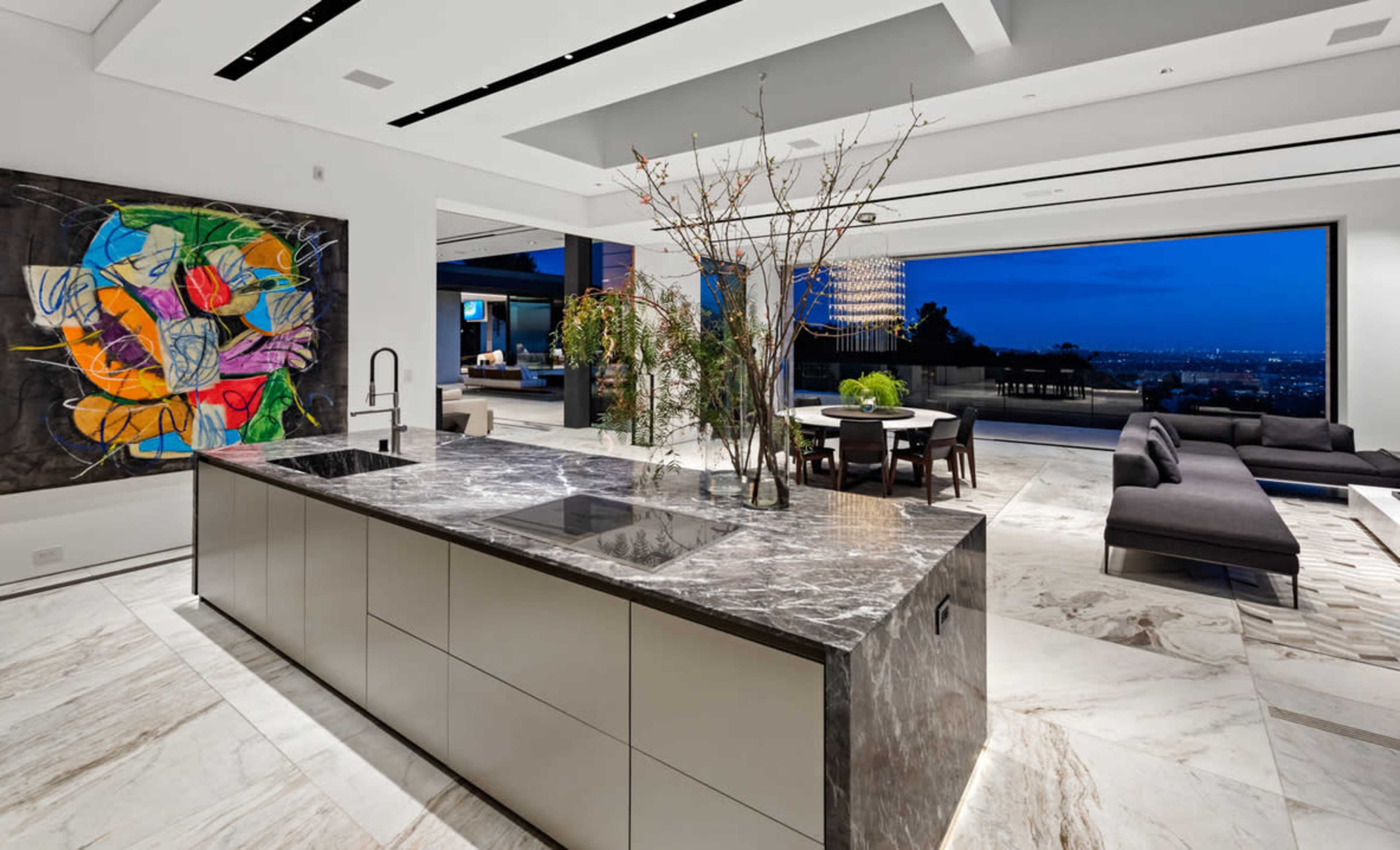 A modern kitchen with a marble island, contemporary decor, and large windows overlooking a city landscape at dusk.