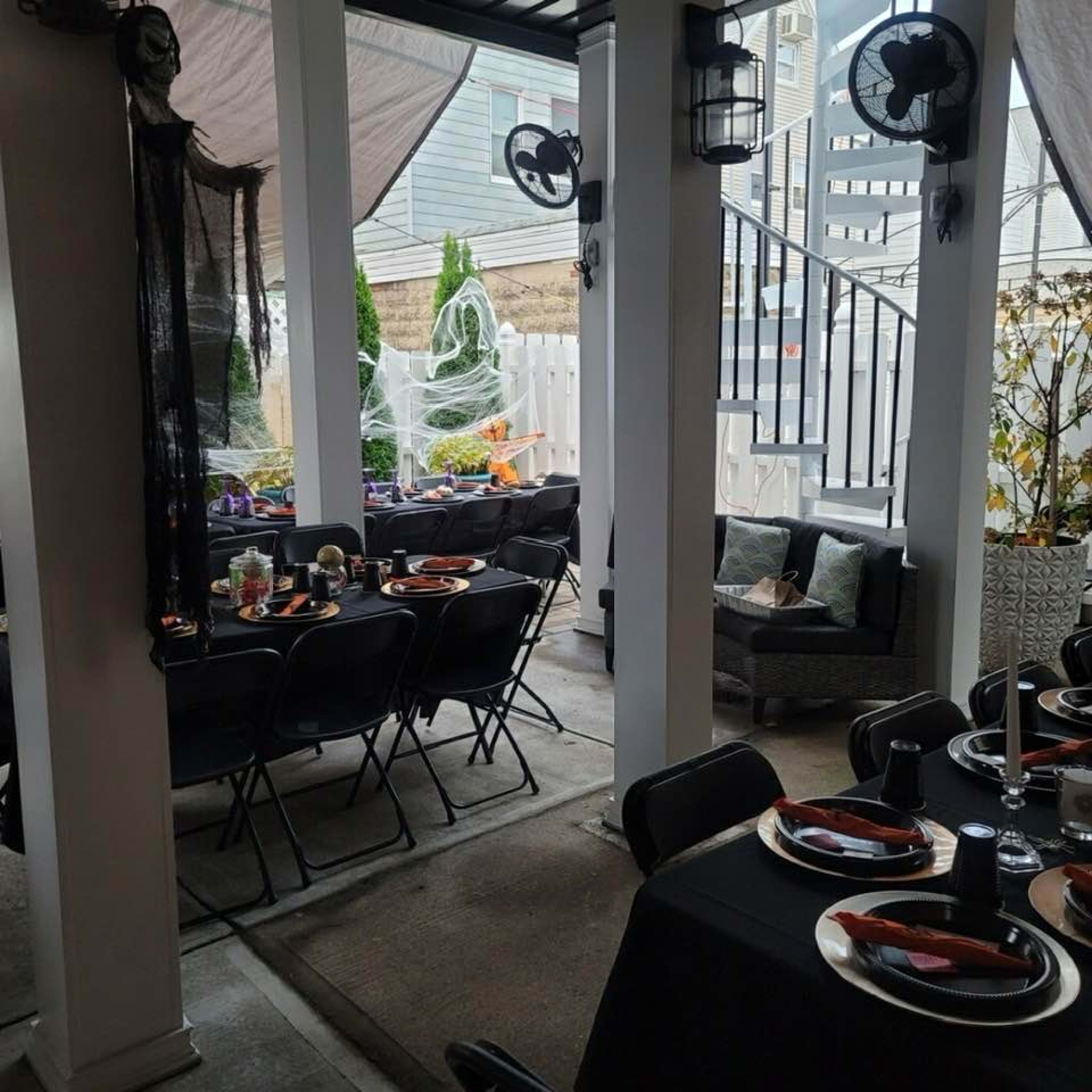 The image shows a decorated outdoor seating area with black tables set for dinner, surrounded by Halloween-themed decorations and a visible staircase in the background.