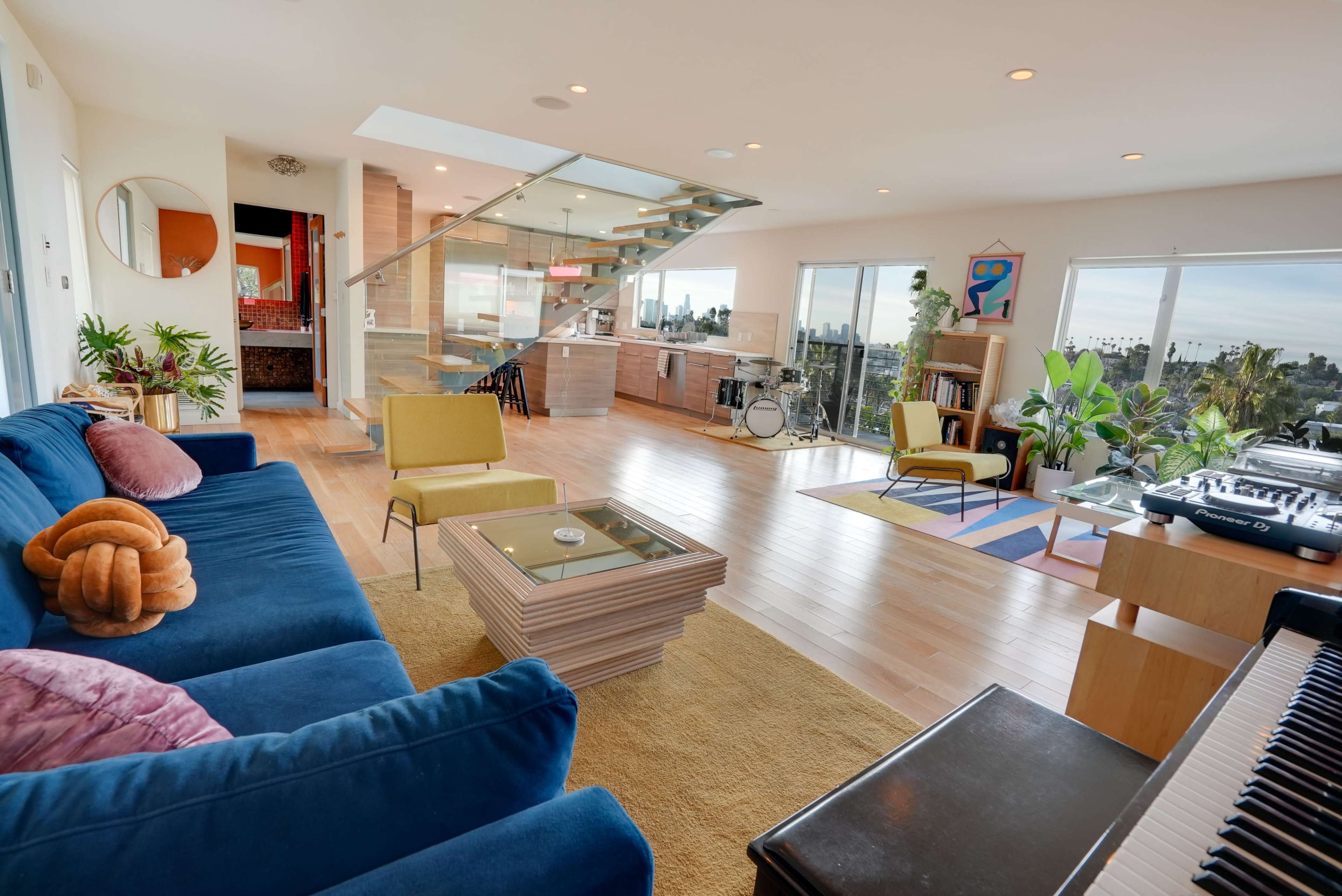 The brightly lit living room features a blue sofa, yellow chairs, a modern coffee table, and large windows showcasing a panoramic view.