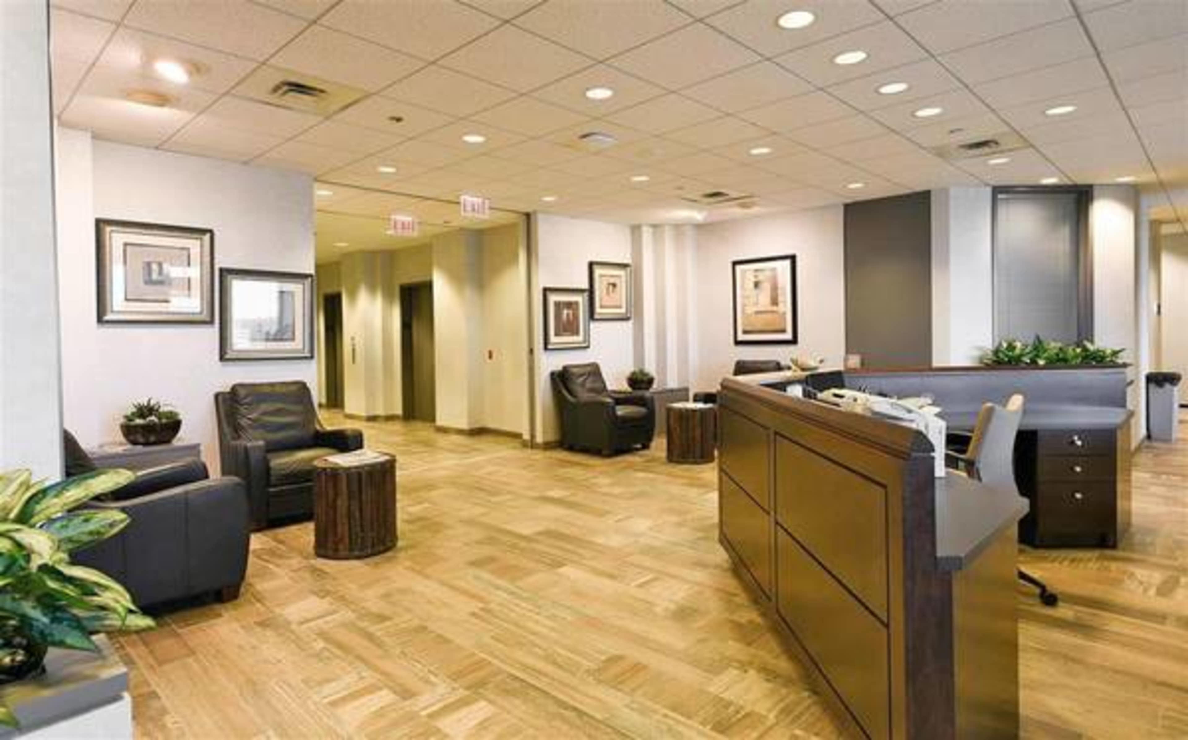 A modern office reception area with two black armchairs, wooden side tables, and a reception desk, surrounded by potted plants and framed artwork on the walls.