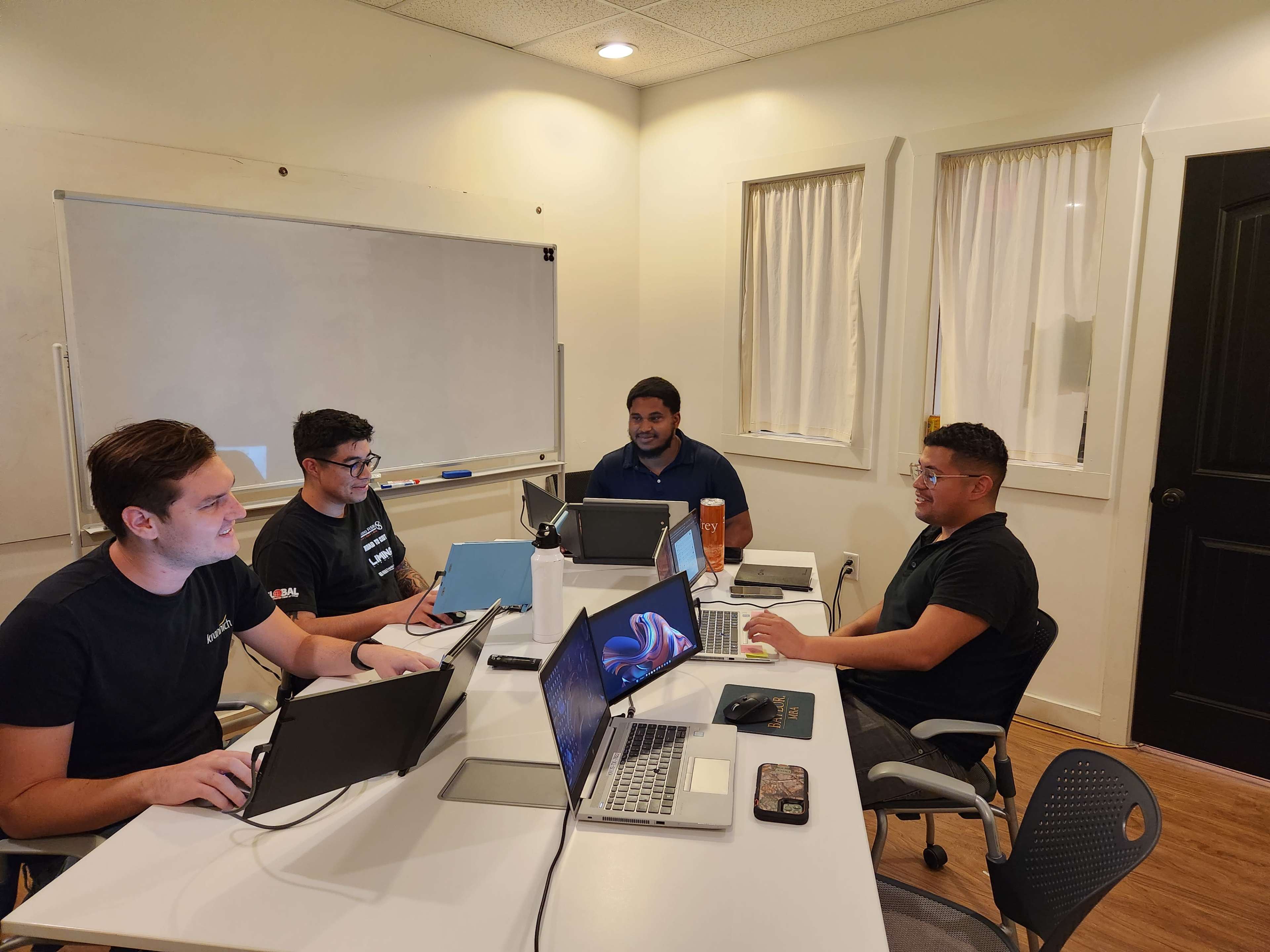 Four individuals are seated at a table in a room with white walls and a whiteboard, working on laptops and discussing ideas.