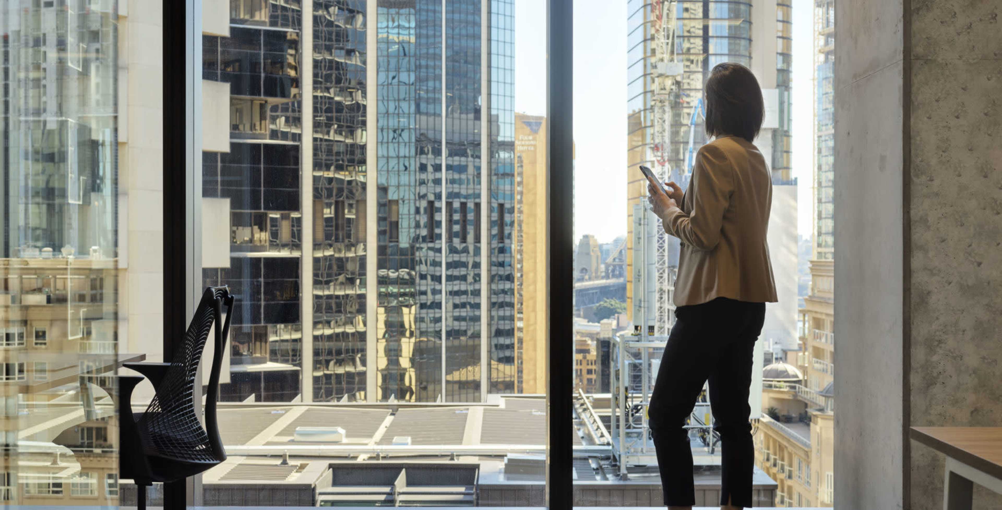 A person stands by a large window in an office, looking out at a cityscape of tall buildings.