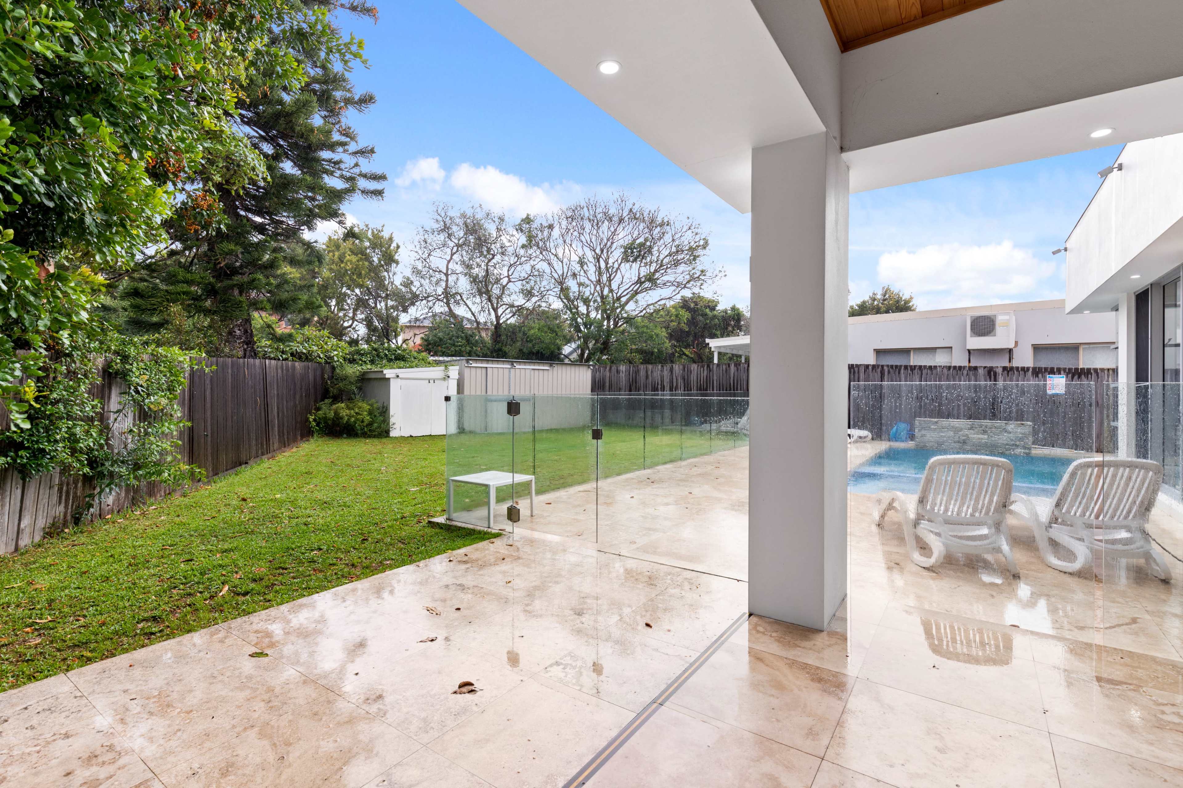 The image shows a modern backyard with a grassy area, a small shed, and a swimming pool, all bordered by a glass enclosure.