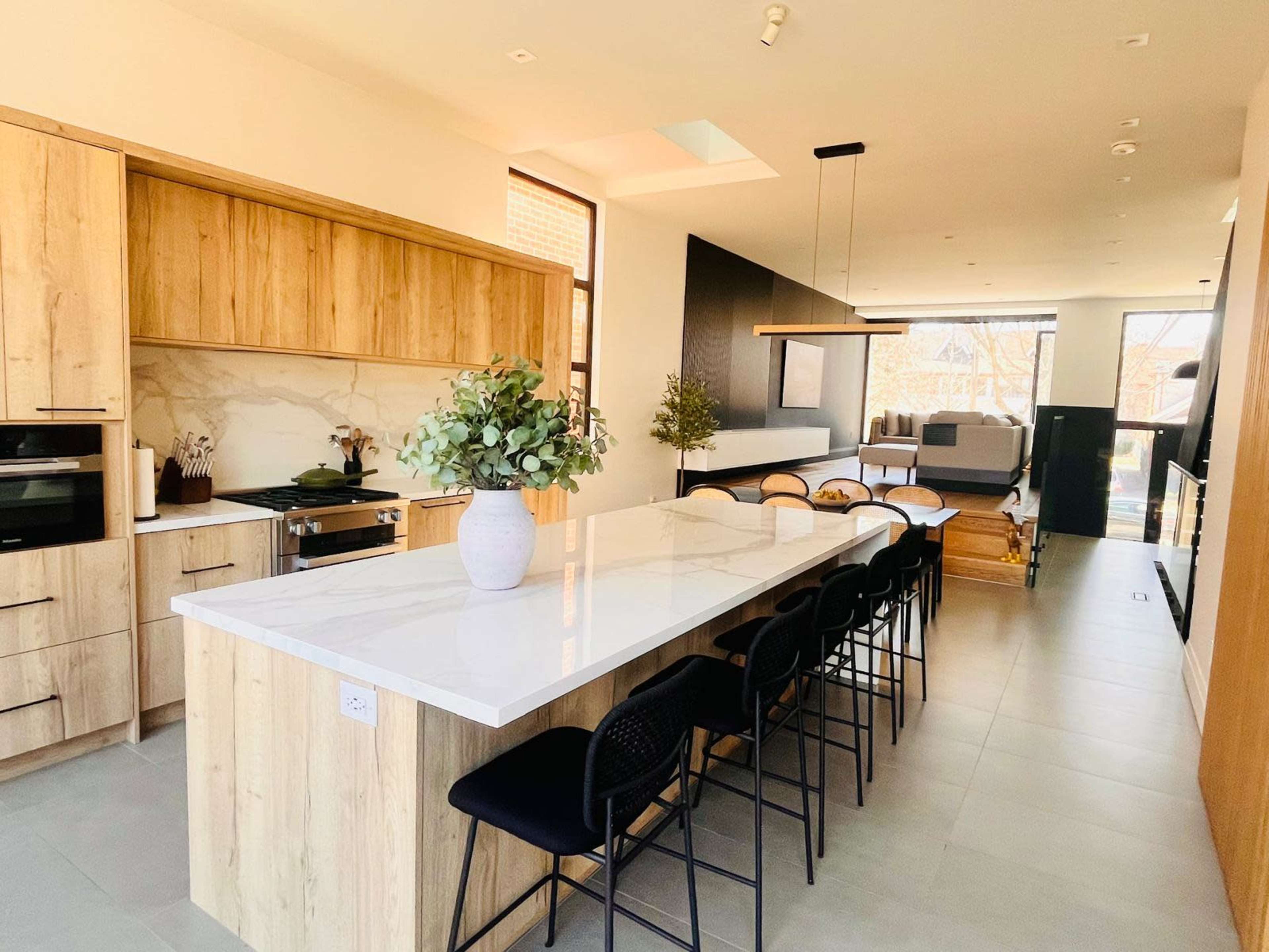 A modern kitchen features a large island with a white countertop, wooden cabinetry, and bar seating, with an open layout extending towards a living area.