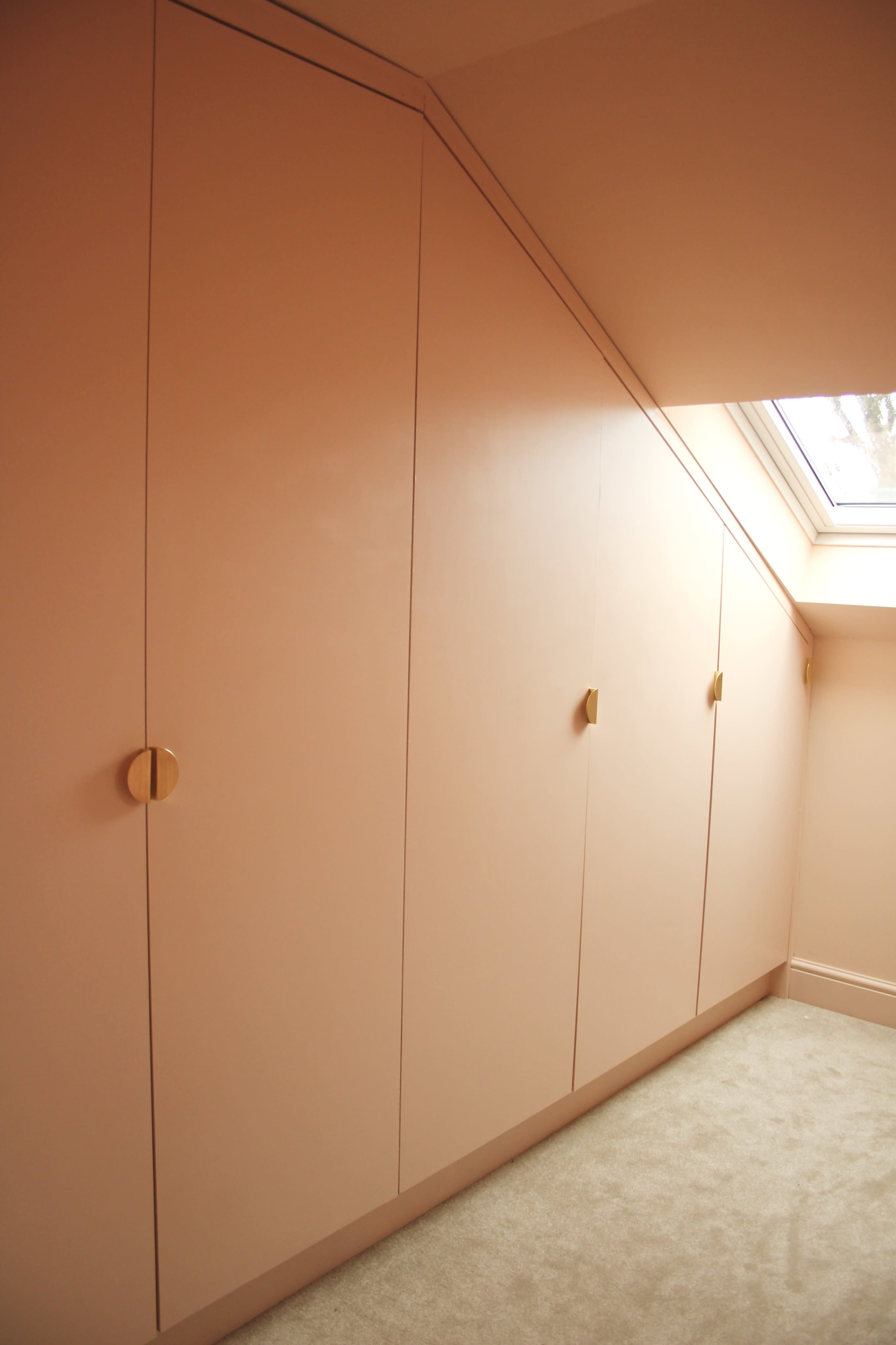 The image shows a series of pink cabinets fitted under an angled ceiling, with gold handles and a skylight above.
