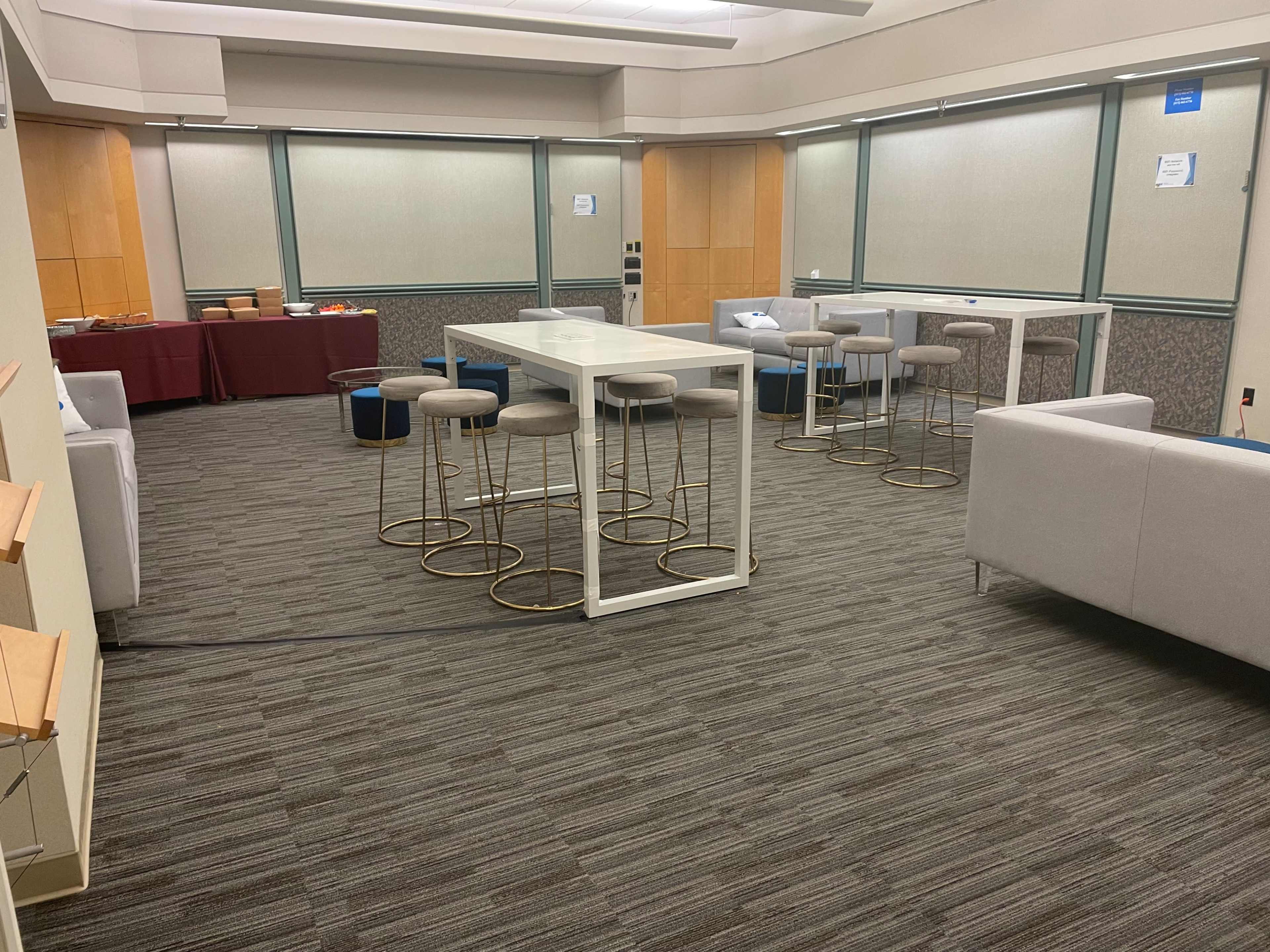 A spacious lounge area with a long table, several bar stools, and seating arrangements, complemented by light-colored walls and carpet flooring.