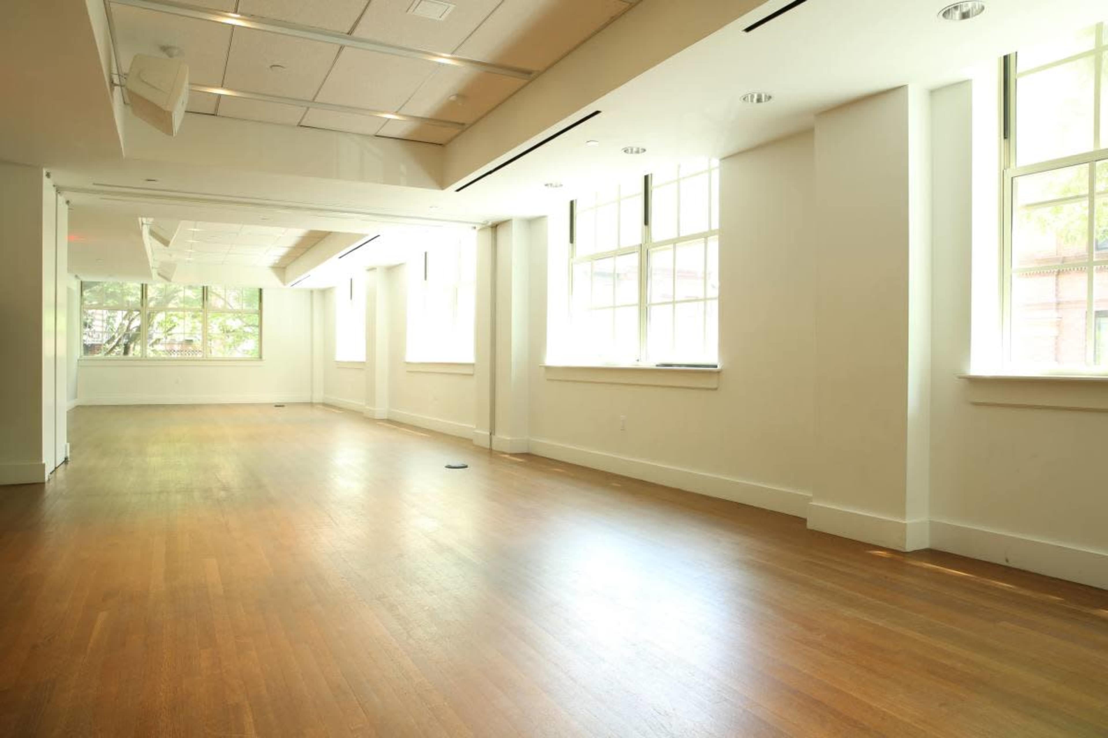 The image shows a spacious, well-lit room with large windows and wooden flooring.