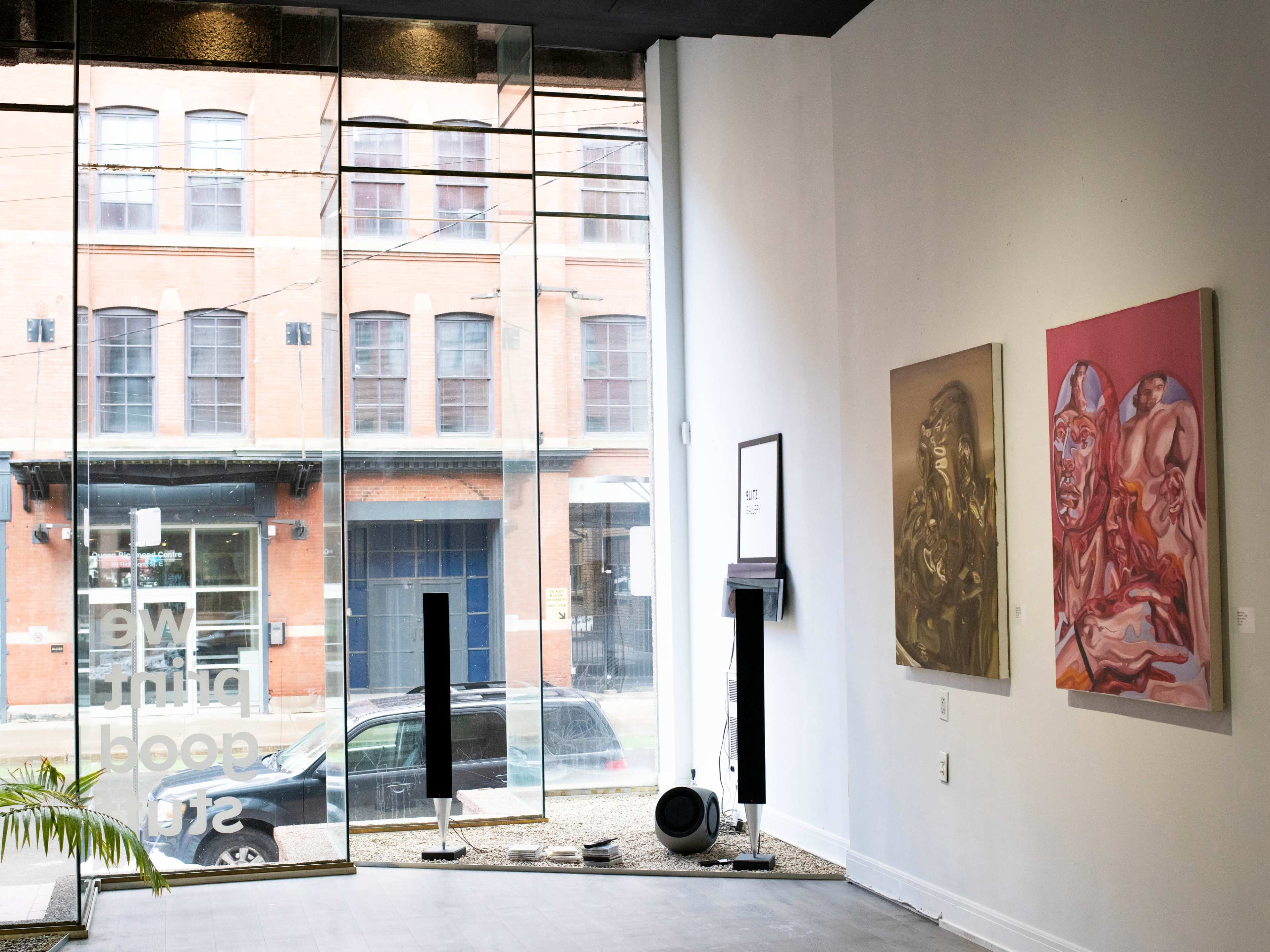 An art gallery with large windows showcasing an urban view, featuring two paintings on the white walls and modern sculptures on the floor.