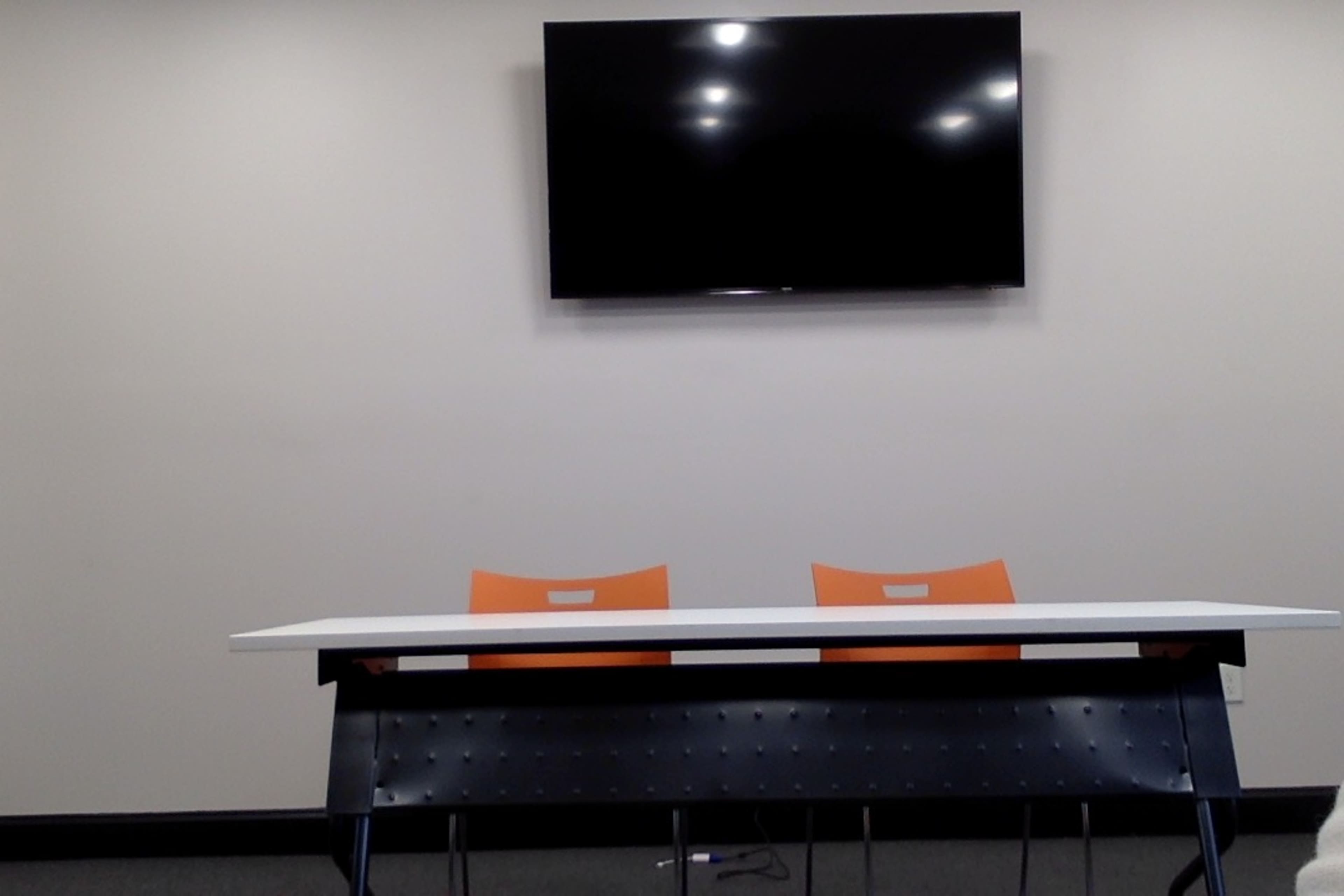 A conference room with a white table, two orange chairs, and a wall-mounted television.