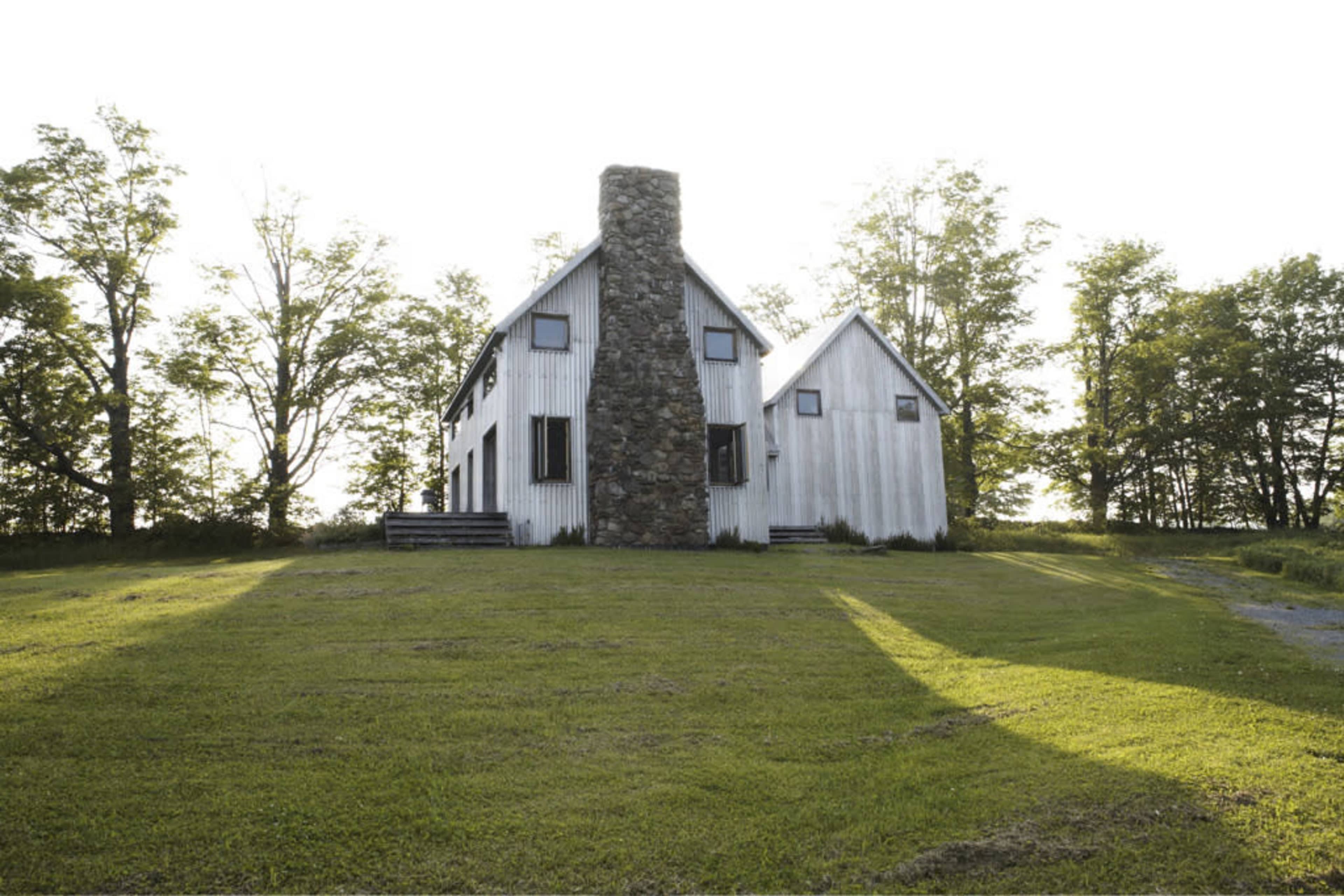 A two-story, white house with a stone chimney stands amidst a grassy area and trees.