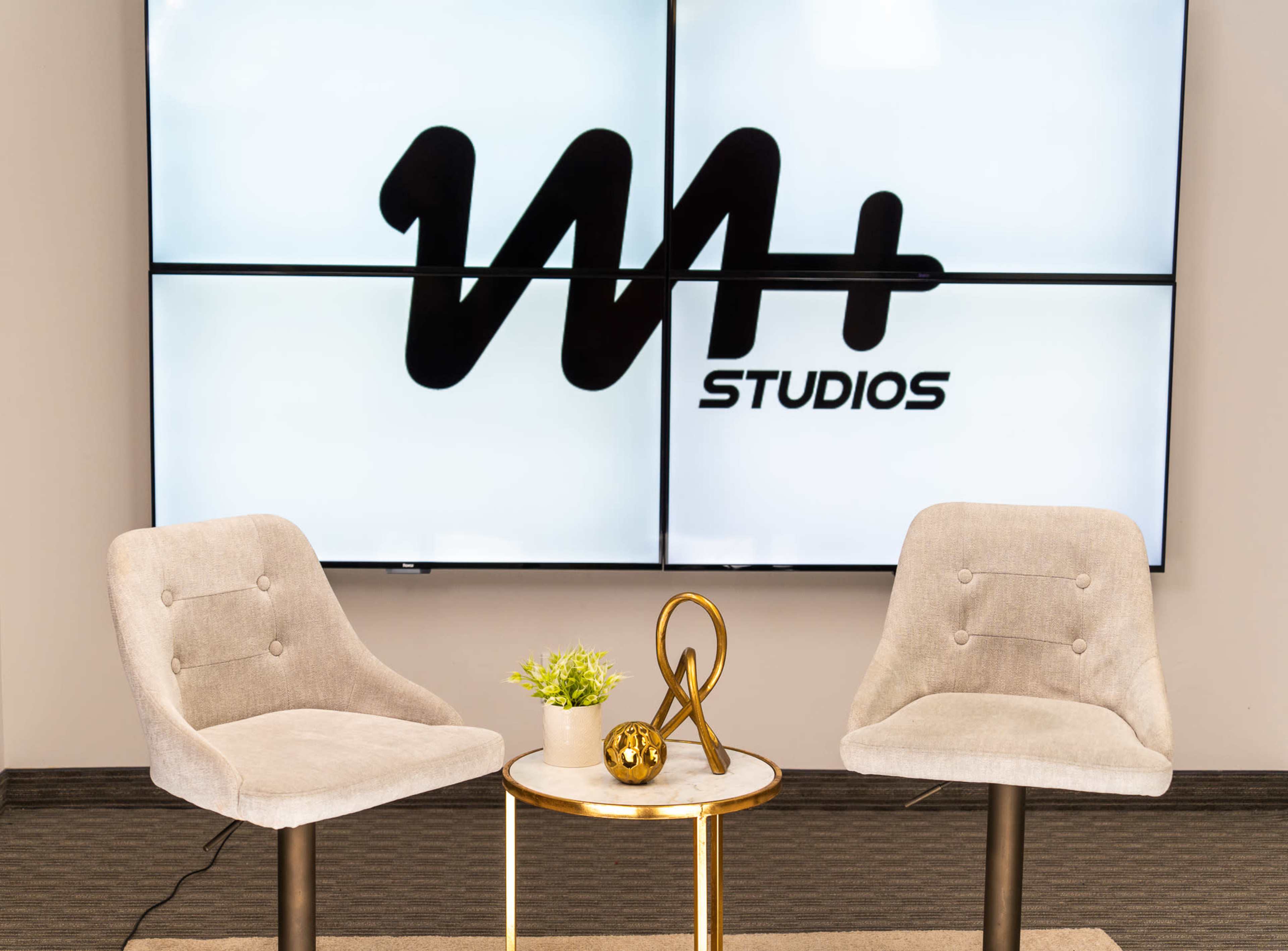 The image shows two gray chairs facing a small round table with decorative items, set against a large screen displaying the logo "W+ STUDIOS."