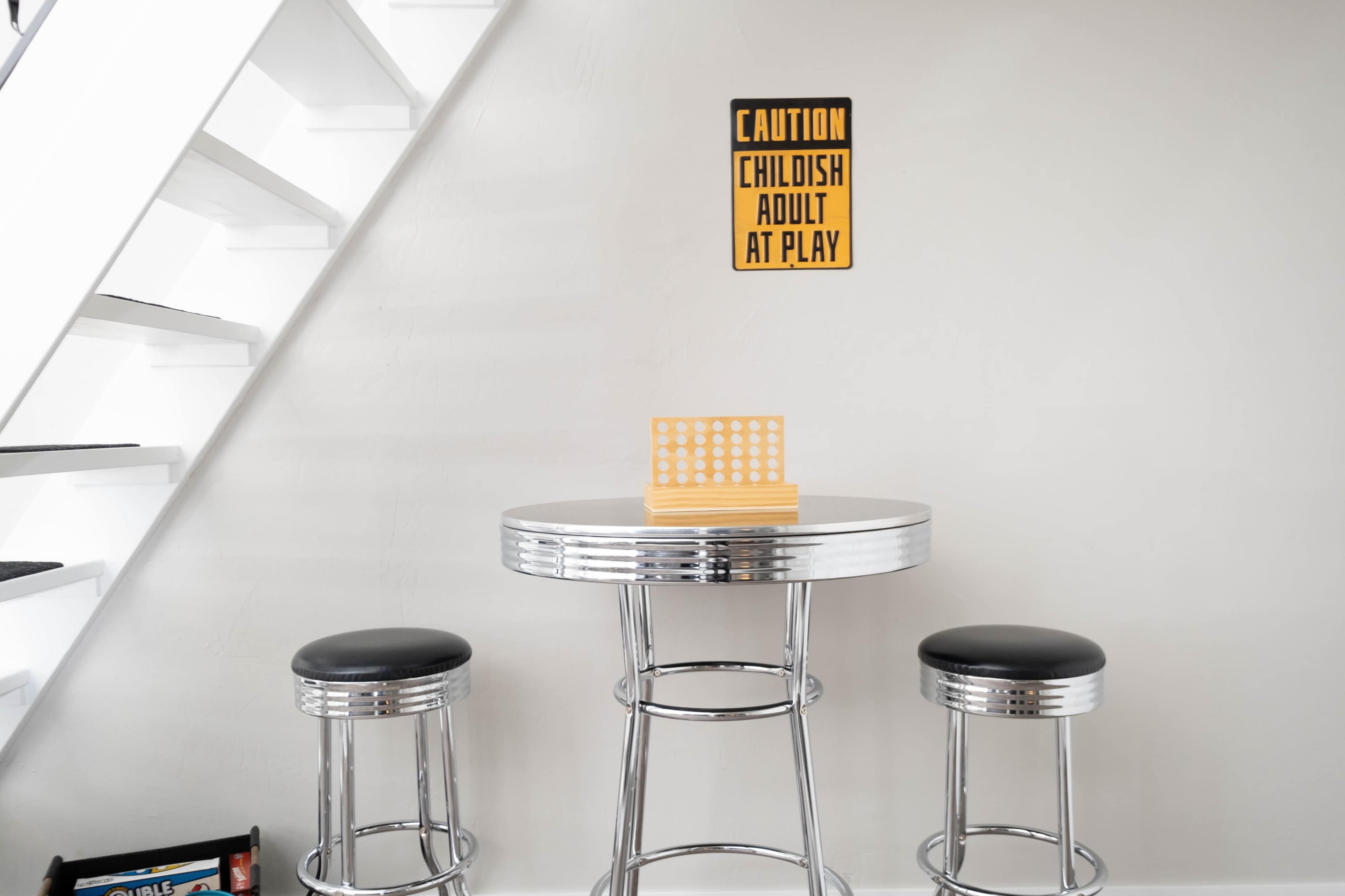 A round chrome table with three black stools sits against a light-colored wall, featuring a sign that reads "CAUTION CHILDISH ADULT AT PLAY" above it.