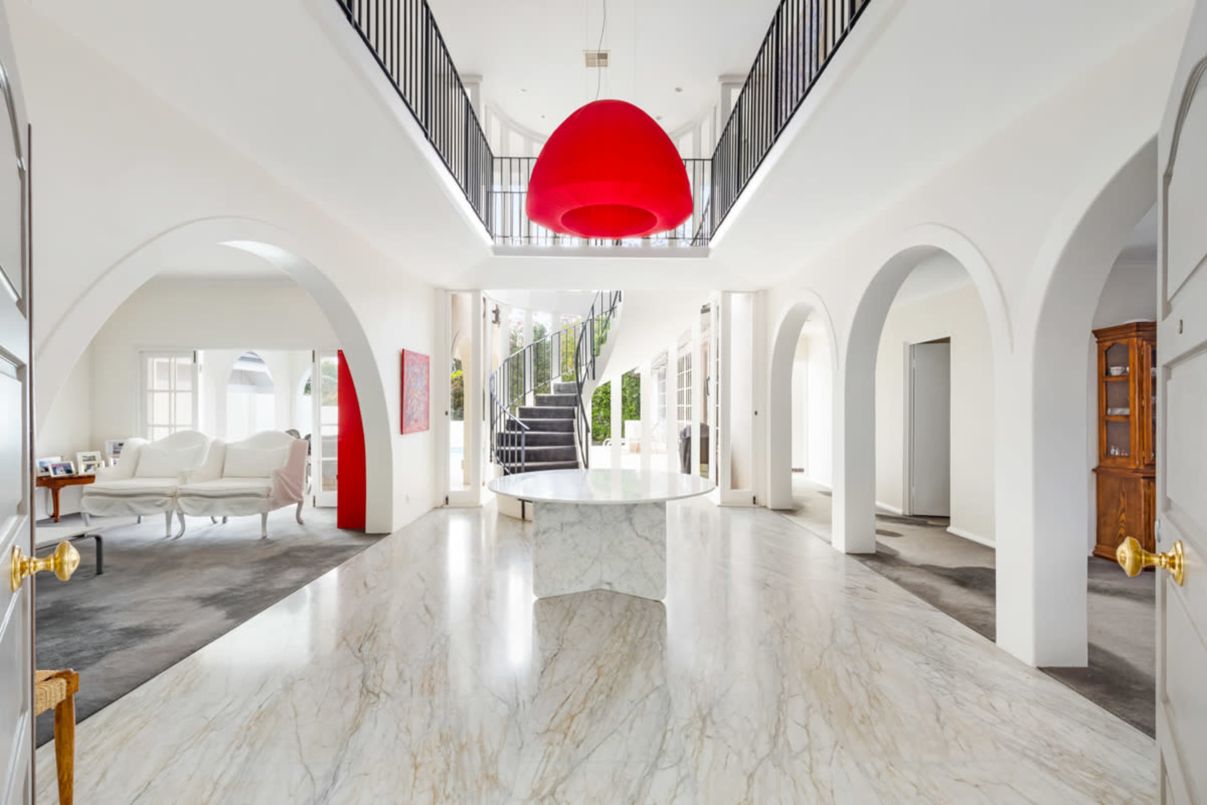 A spacious interior features a large marble floor, arched doorways, a prominent red pendant light, and a sweeping staircase in the background.
