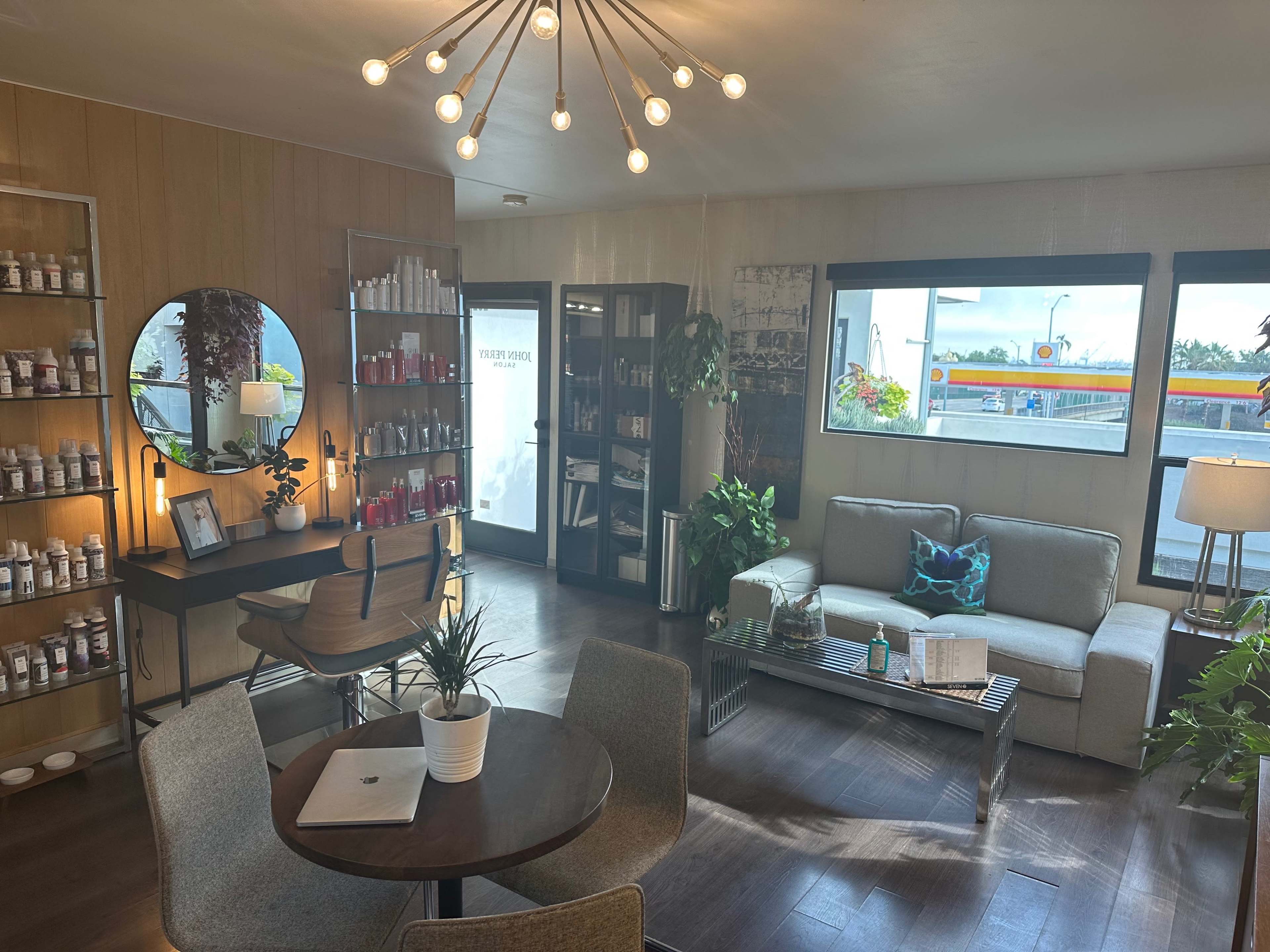 A modern interior space featuring a seating area with a gray sofa, a round mirror, plants, and shelves stocked with products.