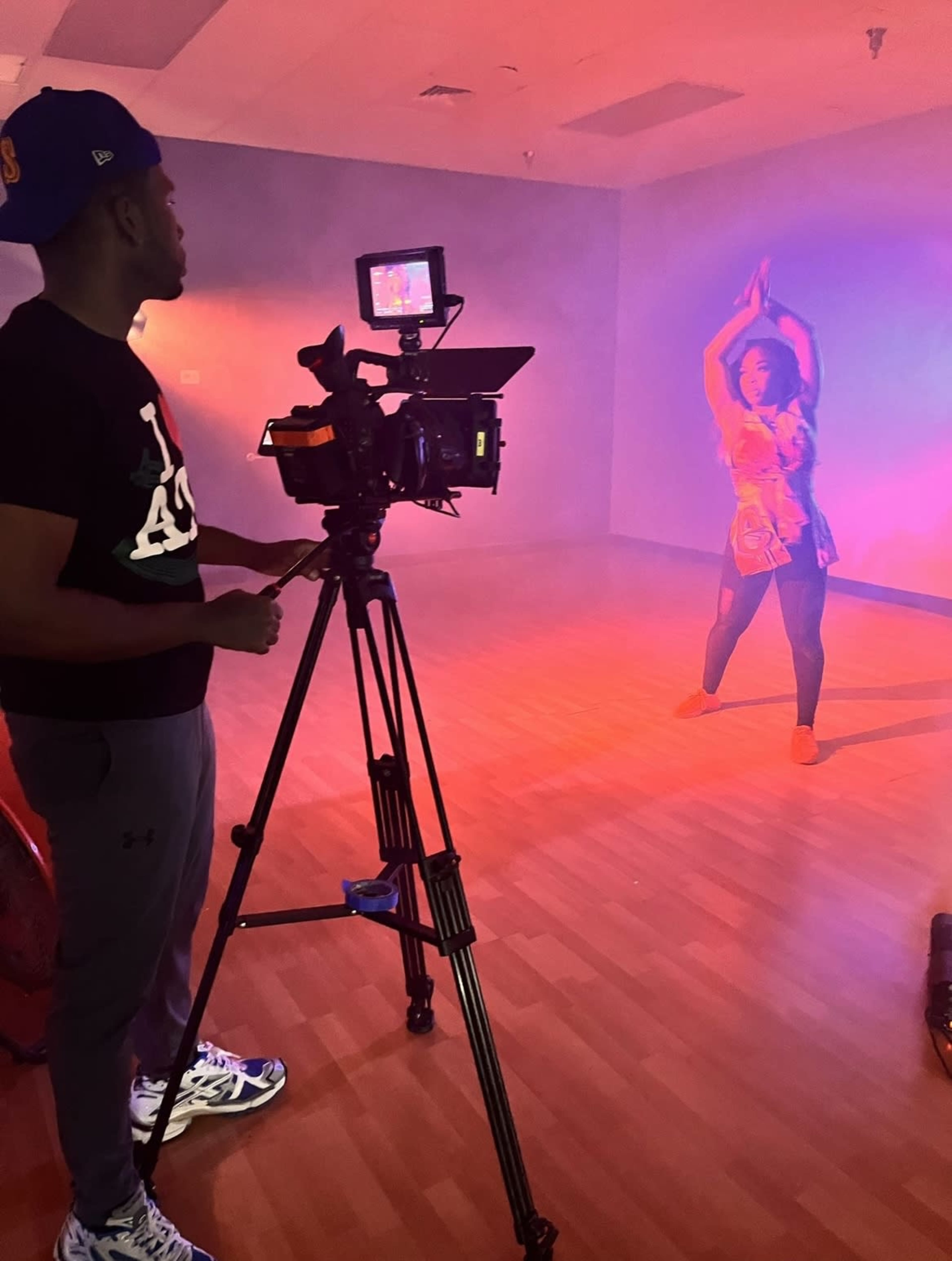 A person operates a camera while a dancer performs in a dimly lit studio filled with colorful lighting.