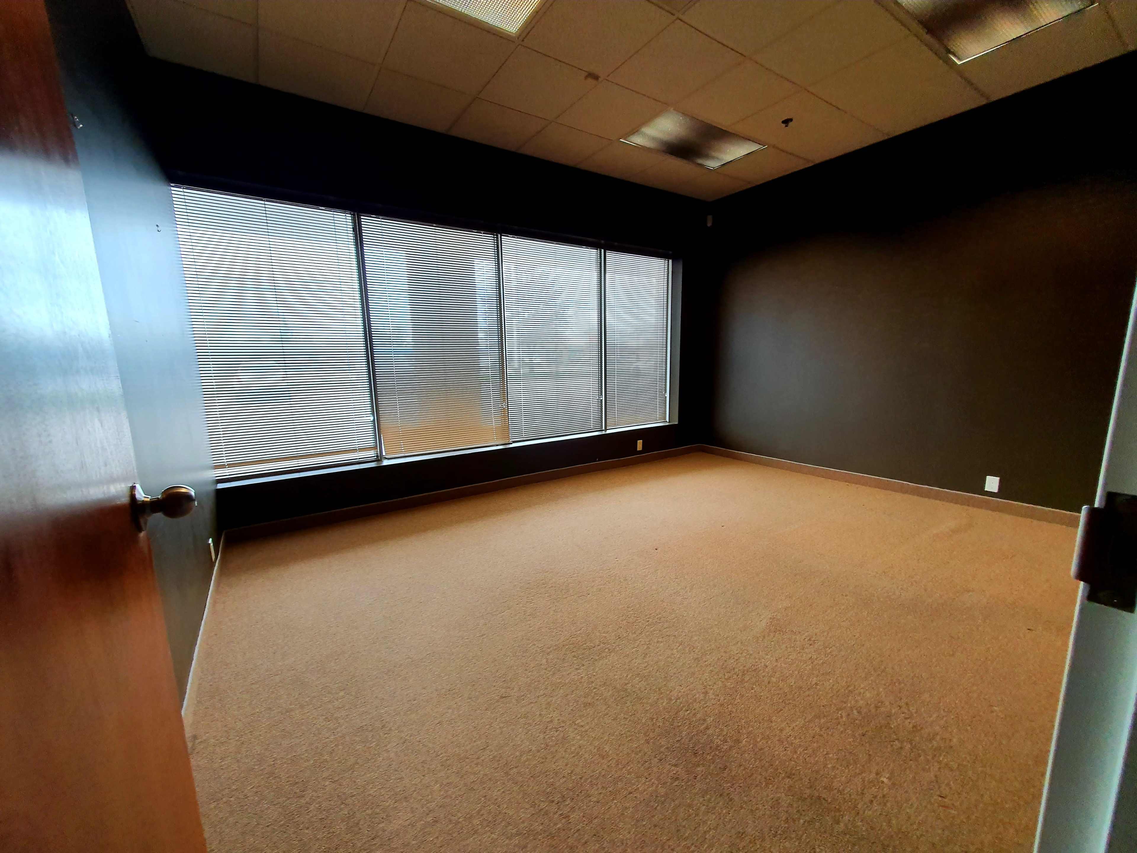 An empty office room with large windows and carpeted flooring.