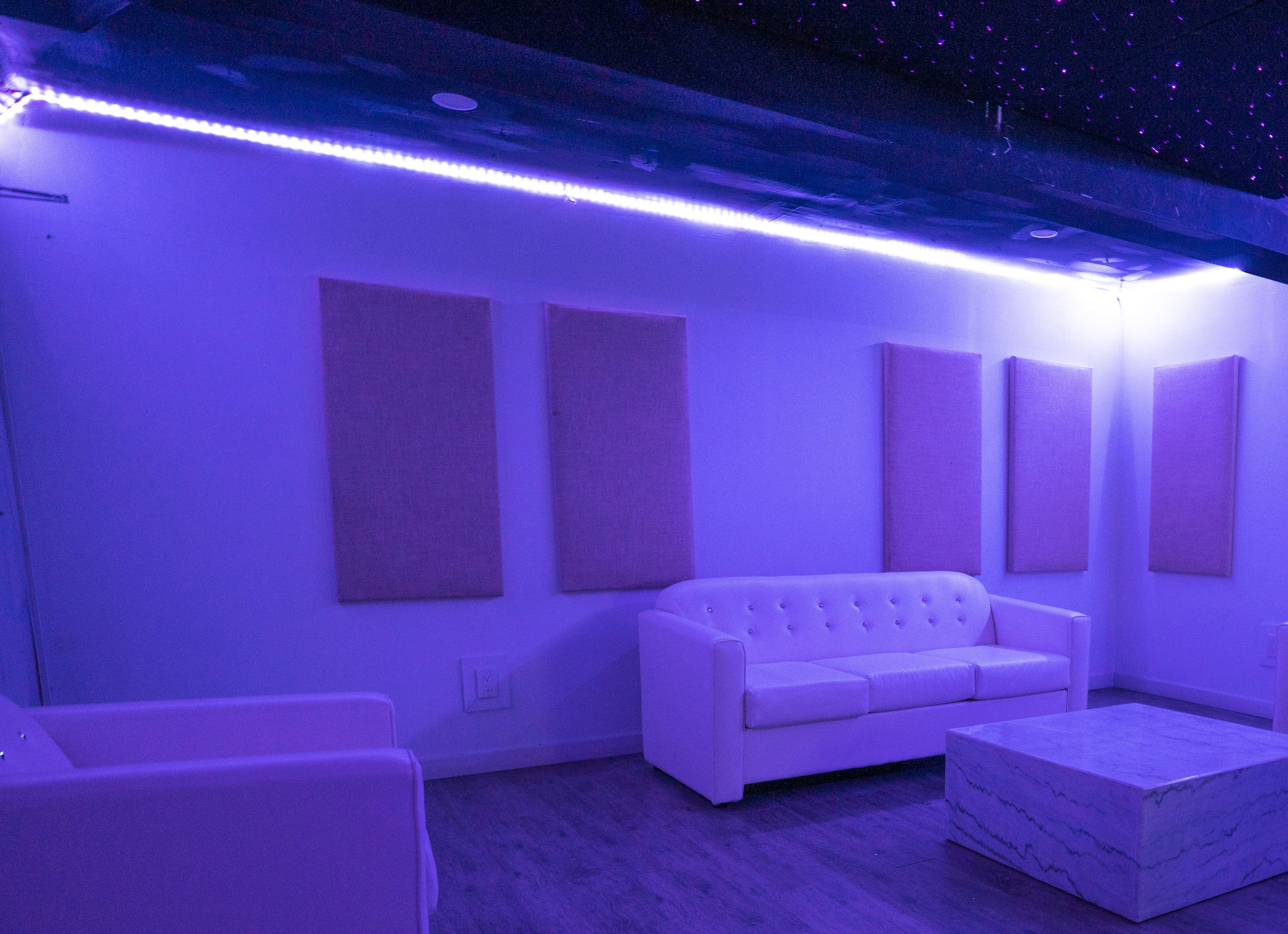 A lounge area with white furniture including a tufted couch and a marble coffee table, illuminated by purple LED lights.