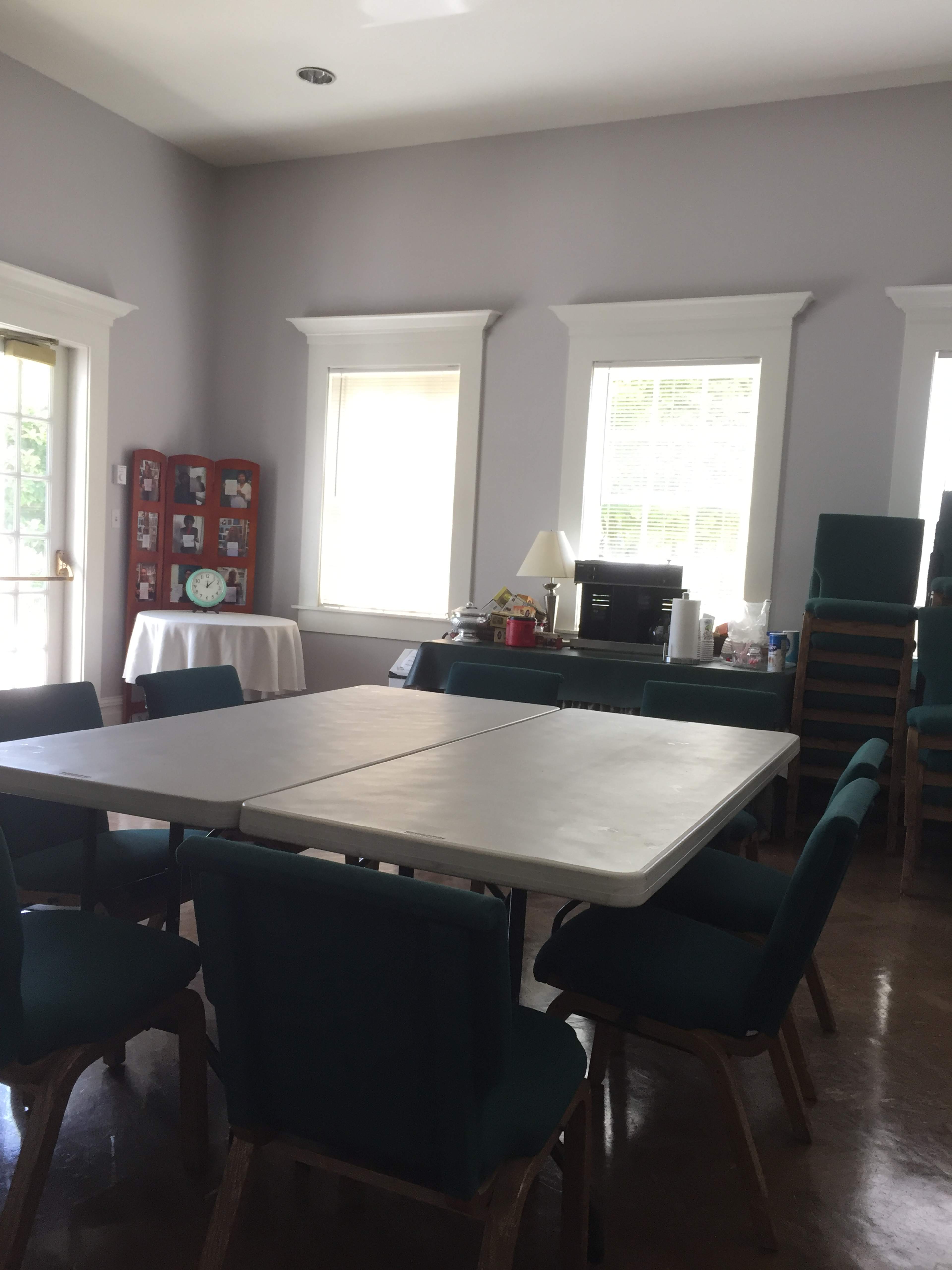 A rectangular table with chairs is set up in a well-lit room featuring windows and a small kitchenette area.