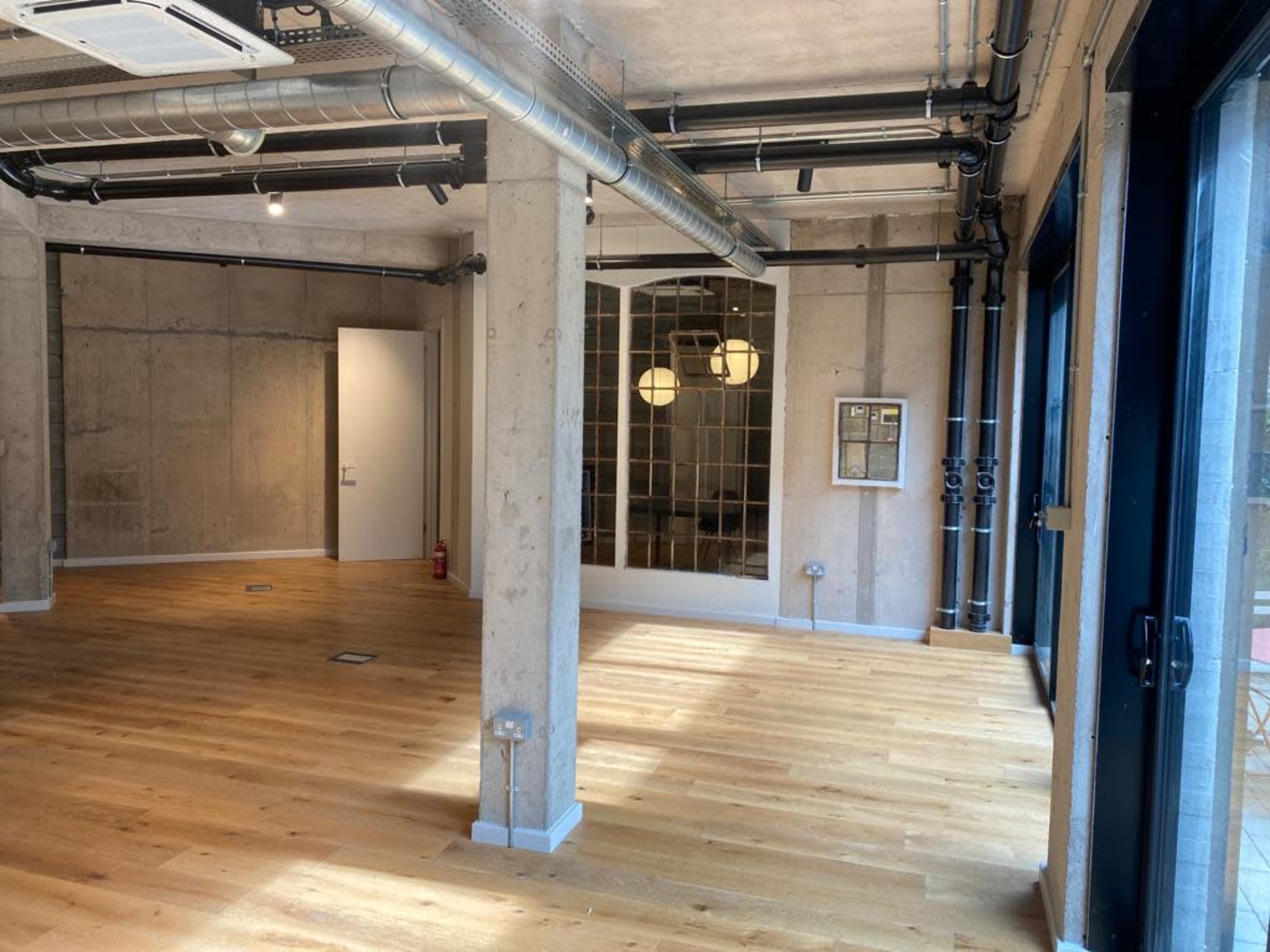 An open office space with concrete walls, wooden flooring, and large windows, featuring industrial-style pipes and minimal furnishings.