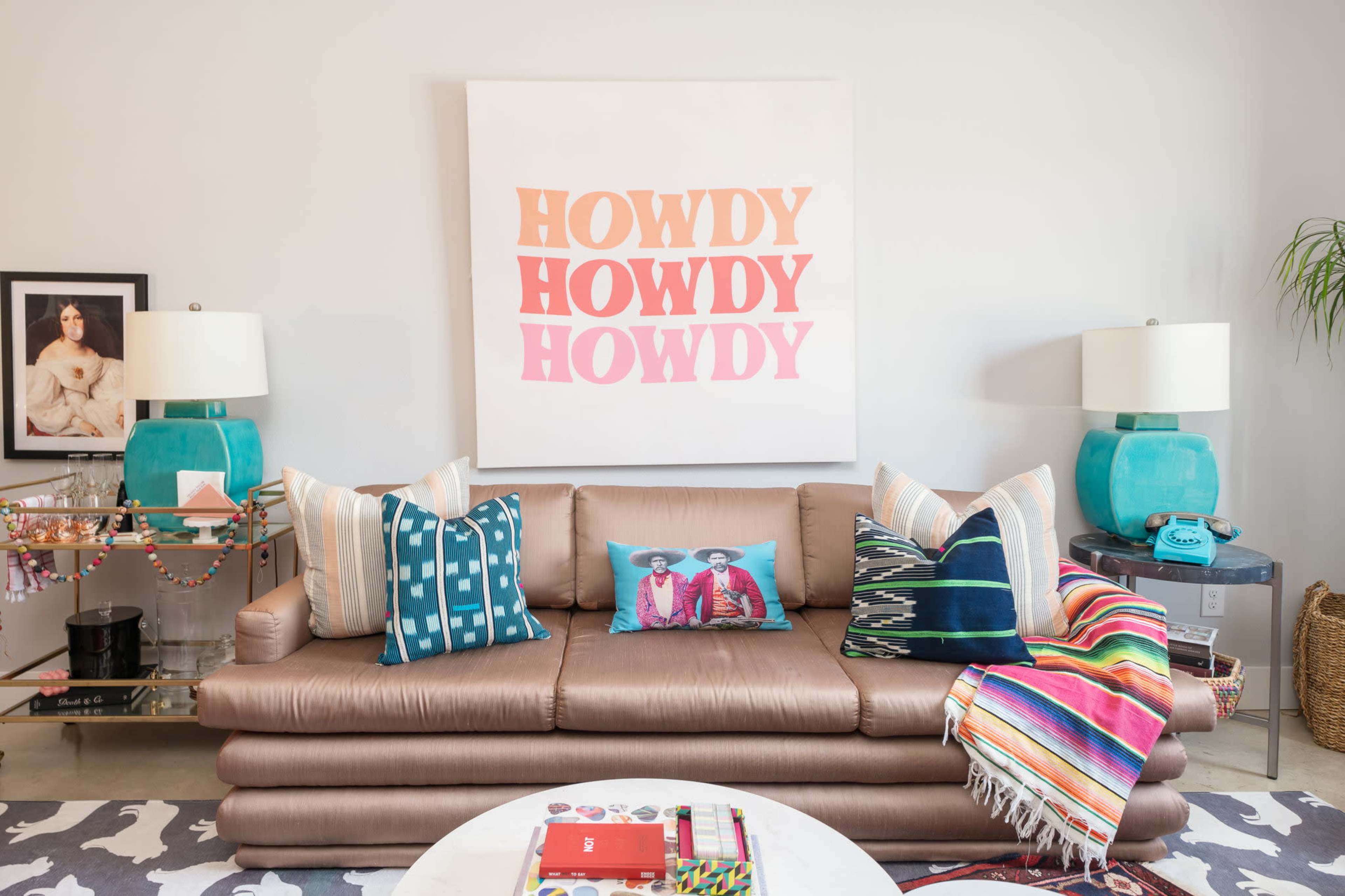 A modern living room features a brown sofa adorned with colorful pillows, a vibrant blanket draped over the side, and a large art piece with the word "HOWDY" displayed prominently above.