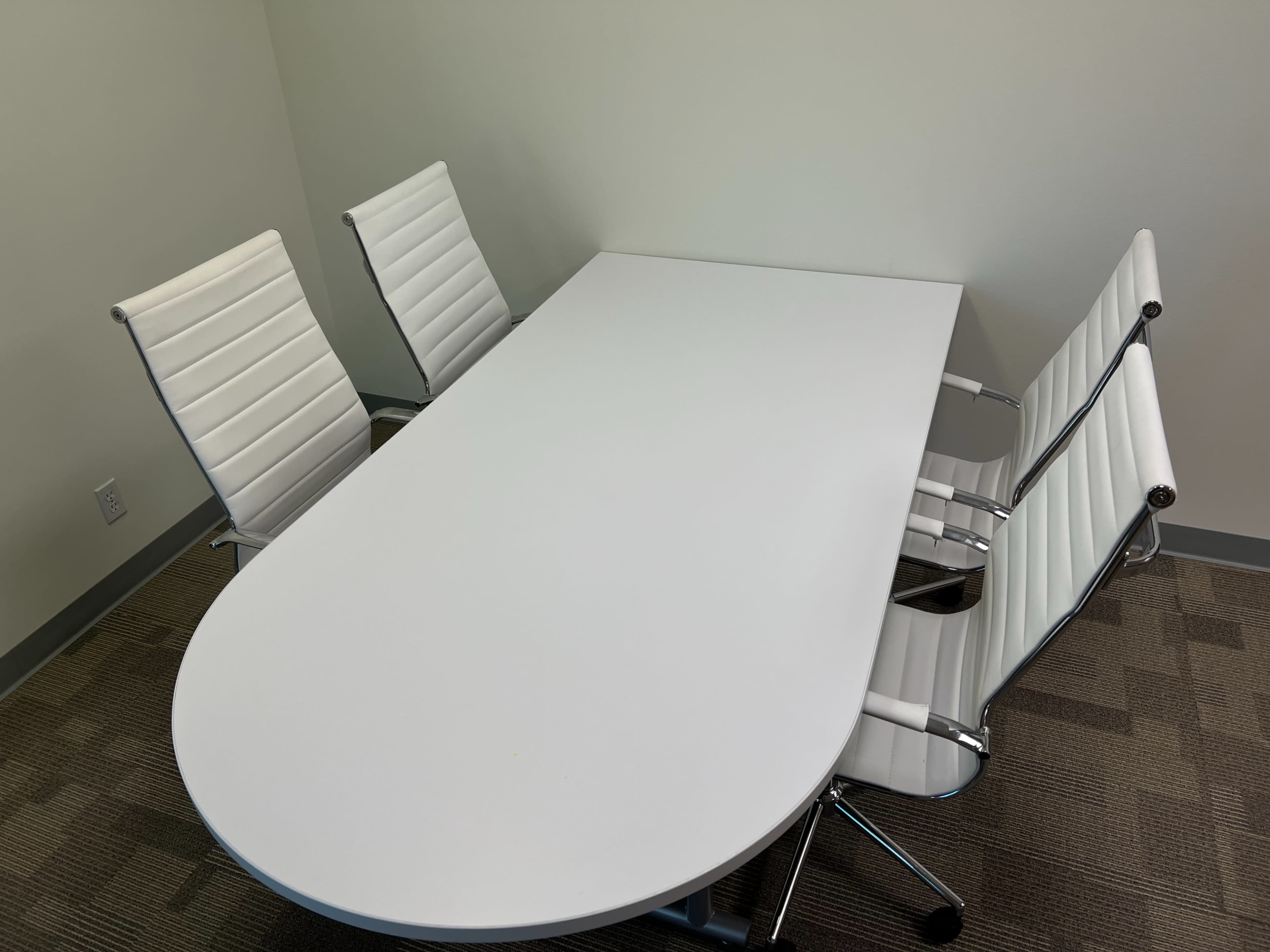A large, oval white table is surrounded by four white chairs in a minimalist conference room.