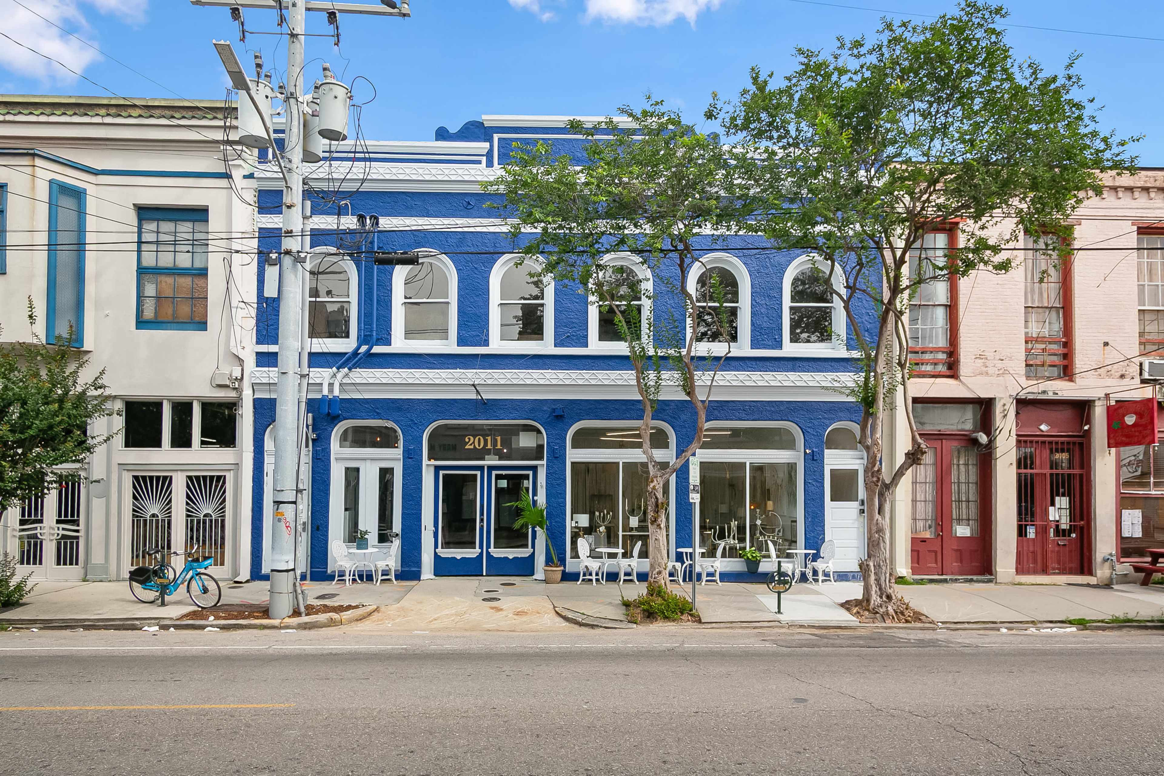 The image shows a blue-painted two-story building with large arched windows and a white awning, flanked by two other buildings in lighter colors, along a street lined with trees and outdoor seating.