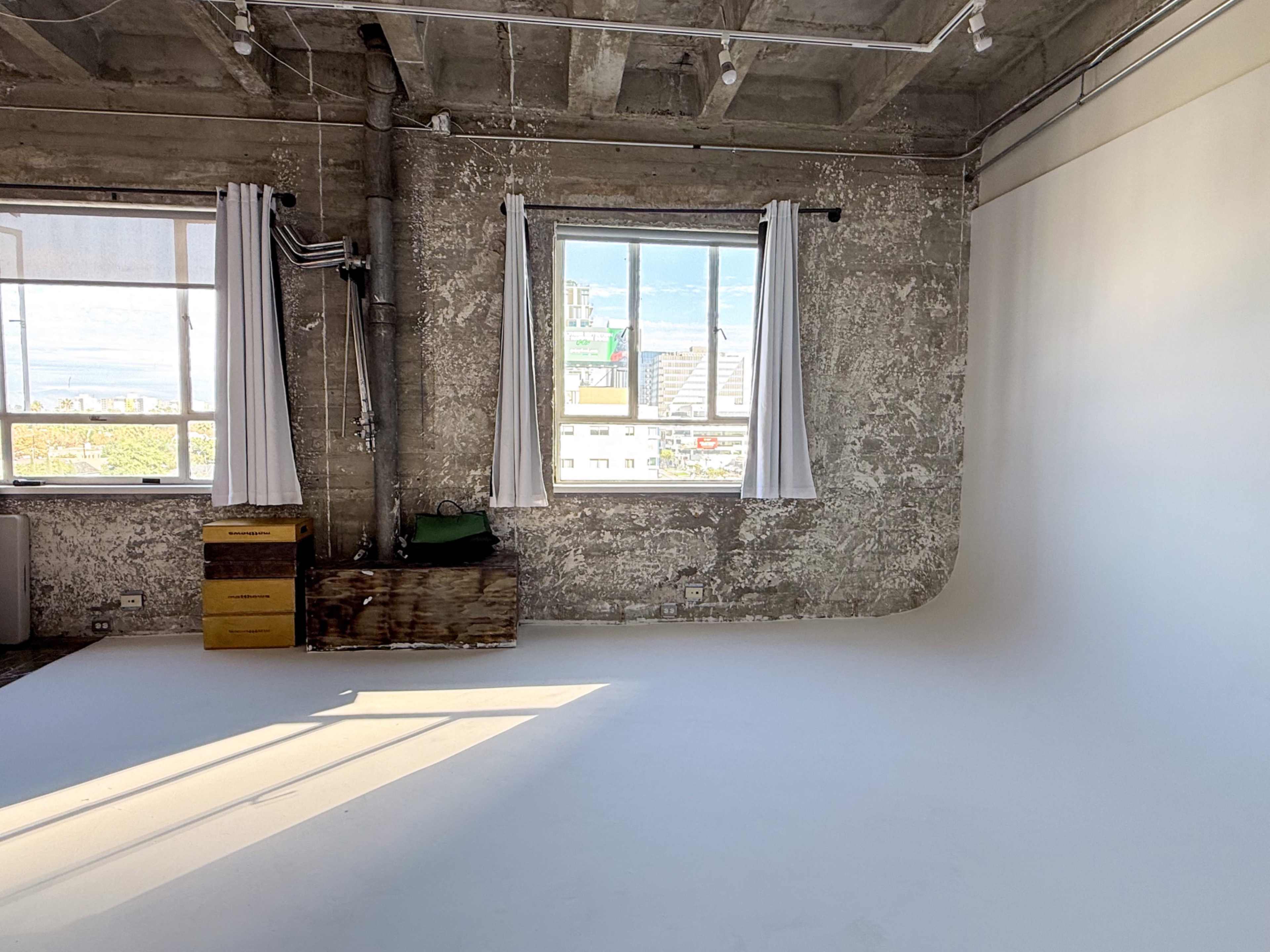 The image shows a spacious industrial-style studio with exposed concrete walls, large windows allowing natural light, and a clean, white backdrop on the floor.