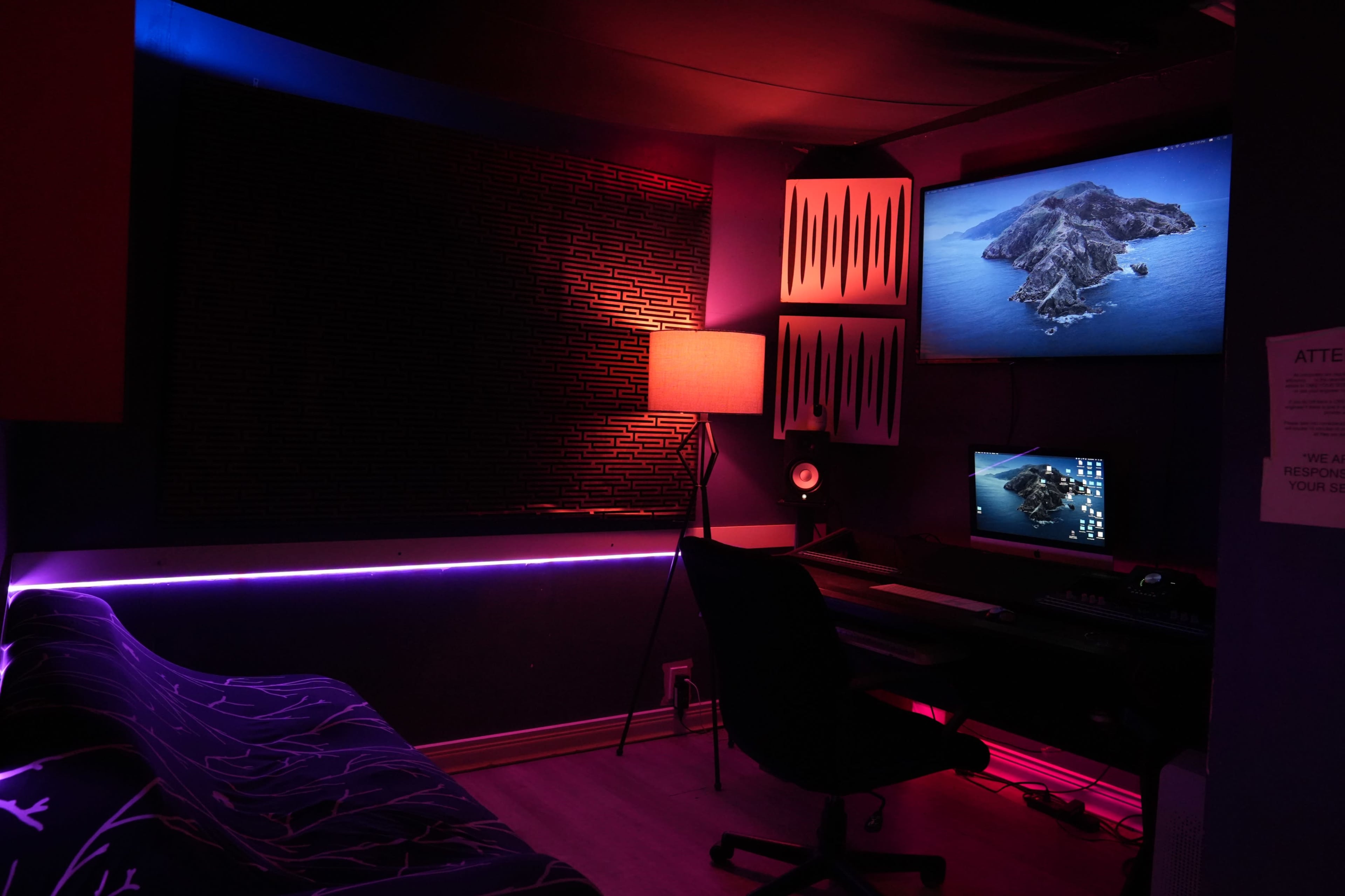 A small, dimly lit room with a desk setup, a computer monitor, and ambient lighting, including a lamp and LED strips.