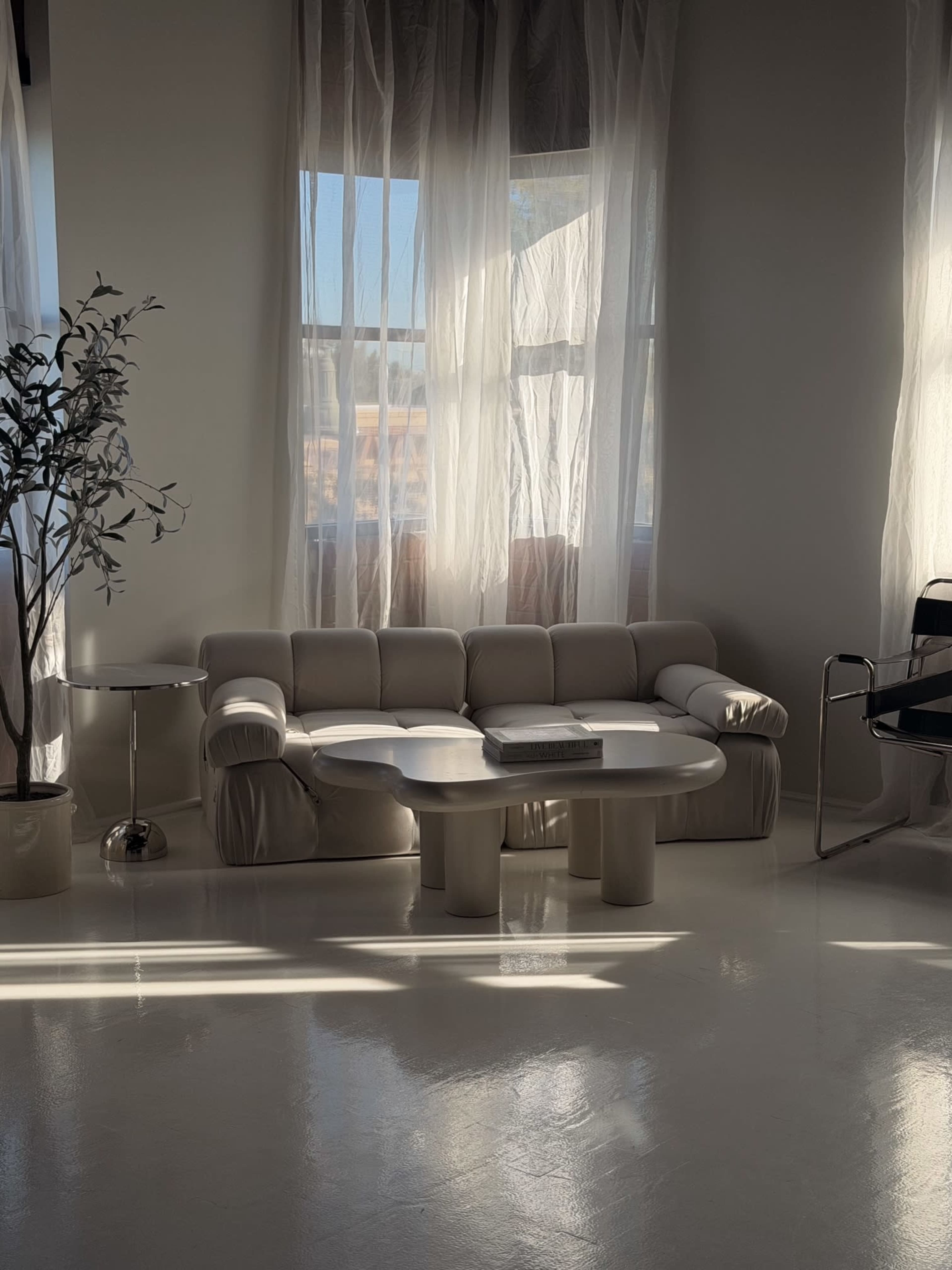 The image shows a minimalist living space featuring a light-colored, sculptural sofa, a round coffee table, and large windows with sheer curtains allowing natural light to filter in.